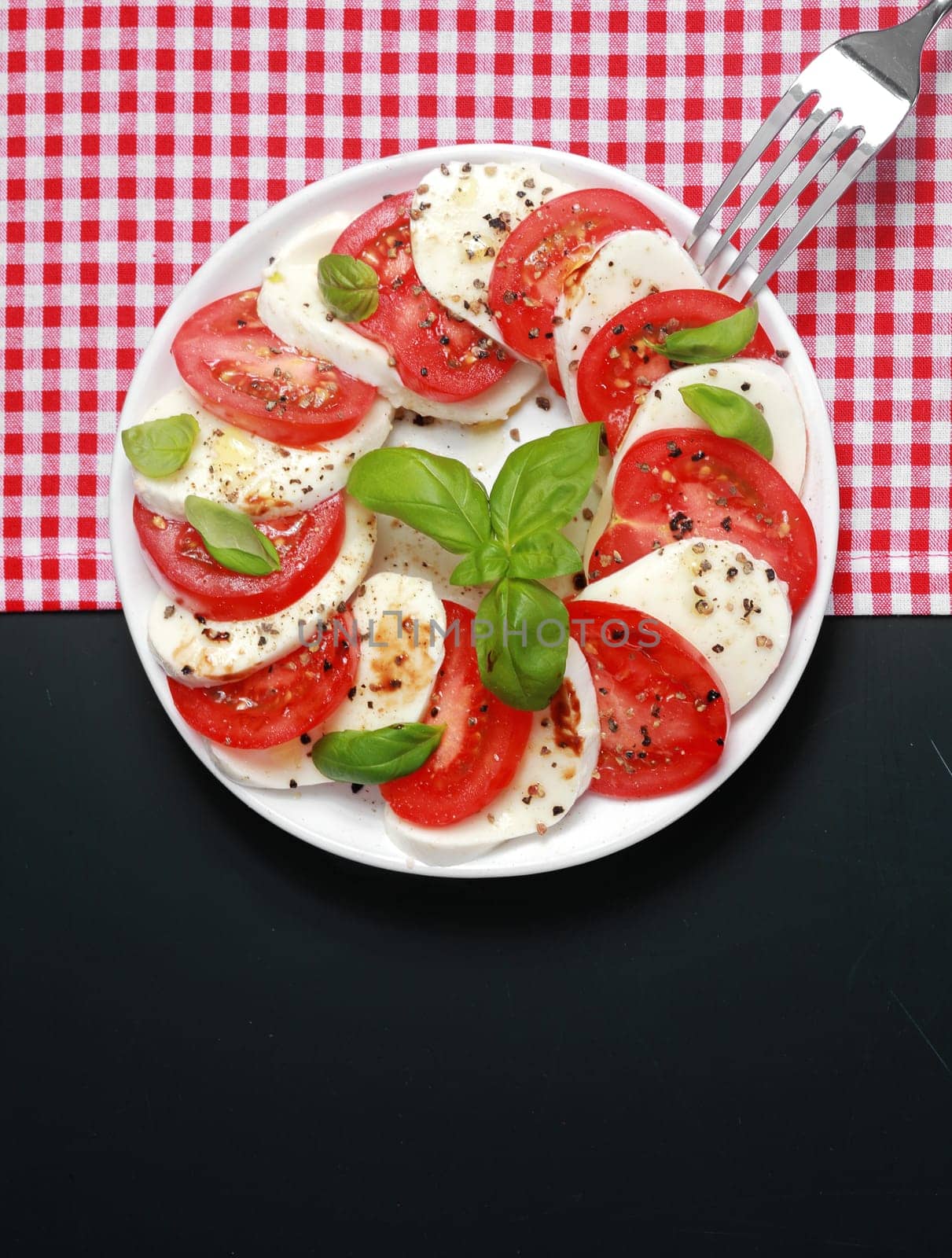 Overhead view of a plate of Italian Caprese salad with alternating slices of tomato and mozzarella cheese seasoned with basil and pepper on a two tone background with red and white check and dark grey