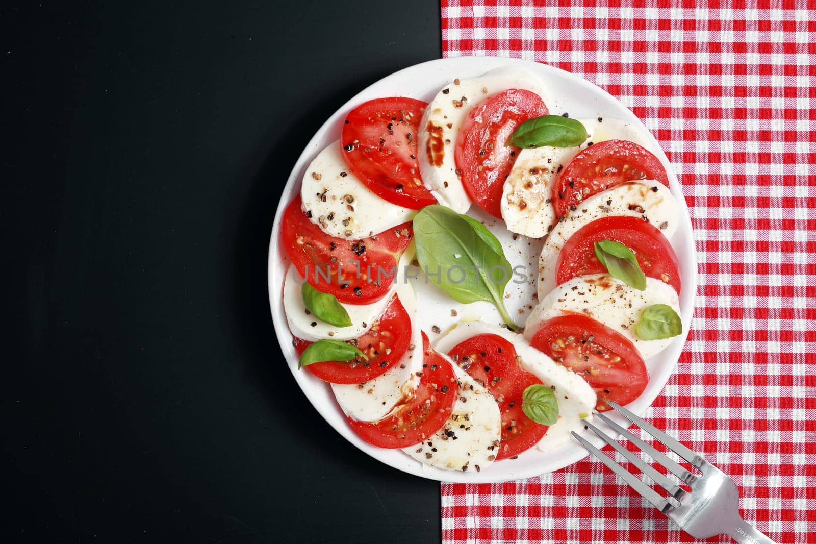 View from above of a plate of Italian Caprese salad with colorful alternating slices of tomato and mozzarella cheese seasoned with basil and pepper on a red and white check and dark grey background