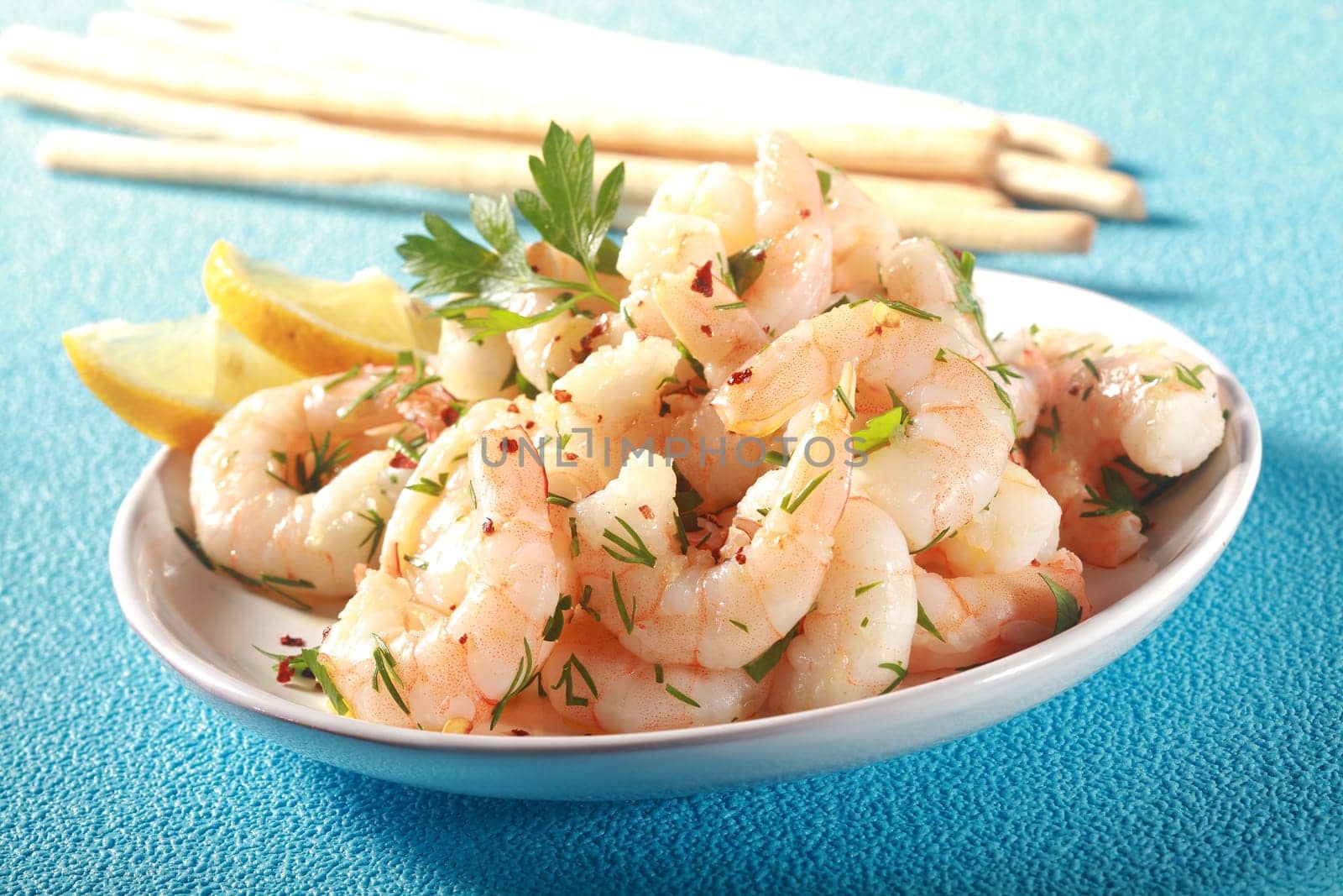 Grilled pink shelled marine prawns seasoned with olive oil and herbs served with lemon and bread sticks for a tasty seafood starter to dinner, on turquoise blue