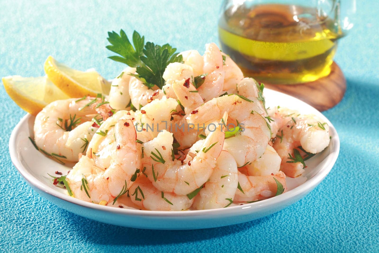 Grilled pink shrimps seasoned with fresh herbs and drizzled with olive oil served in a dish with lemon as a delicious seafood delicacy, on a textured blue table
