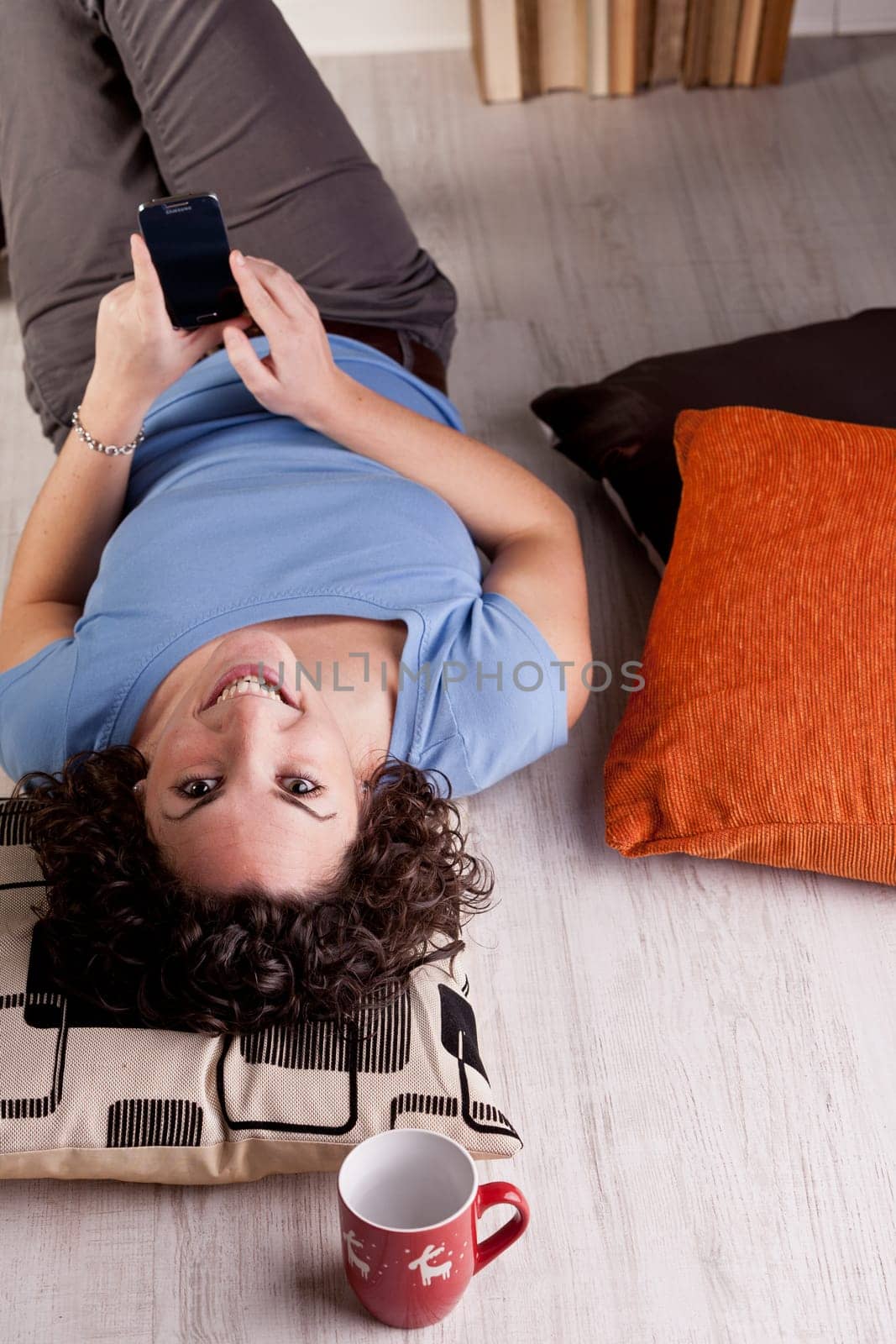 A smiling girl staring at camera with a mobile phone in her hands lying on the floor of her room