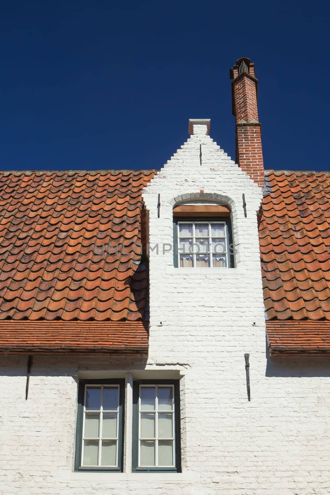White brick house with three windows and red roof. (Bruges, Belgium). Blue sky in the background.