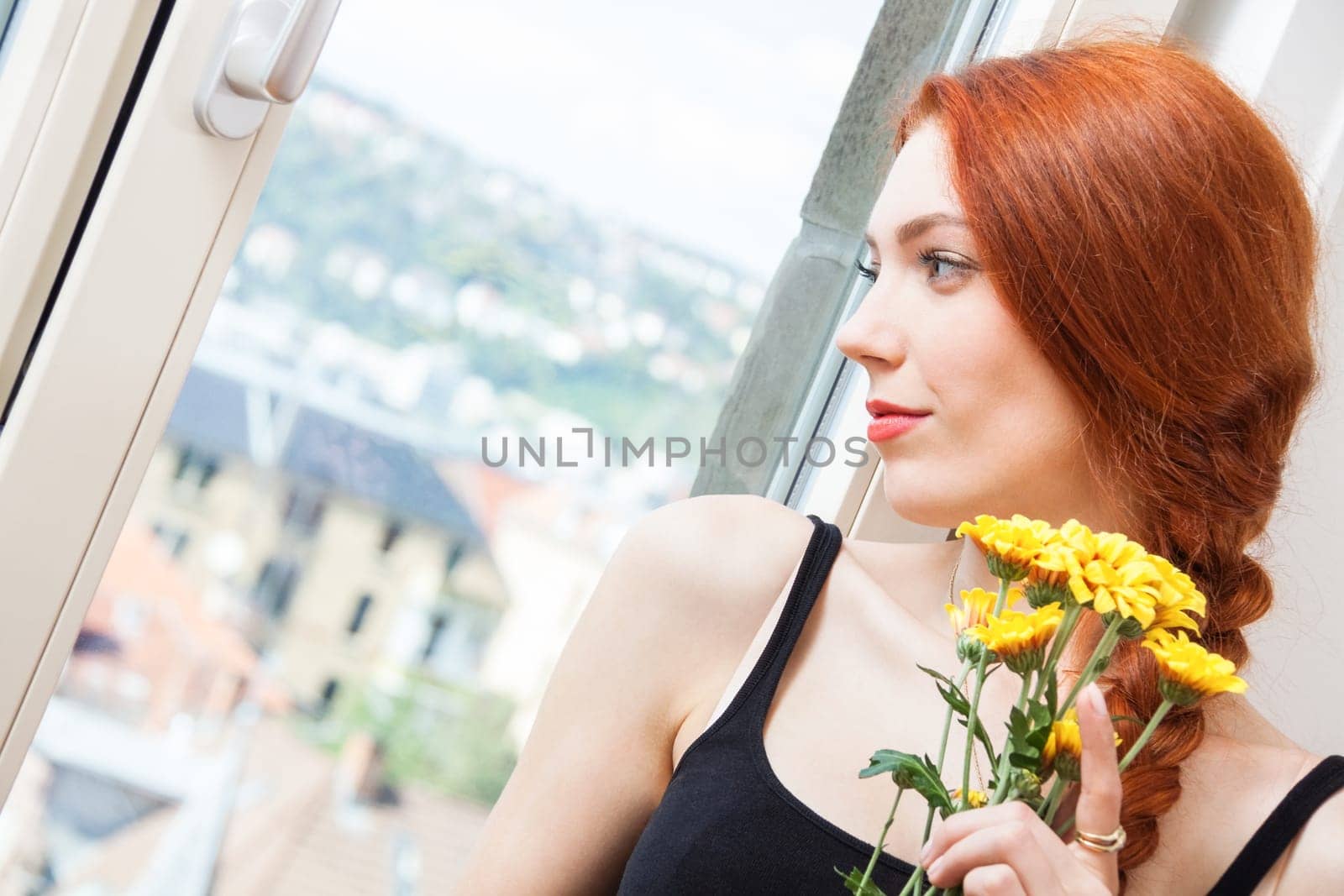 Thoughtful young woman looks out the window in summer with flowers in her hand