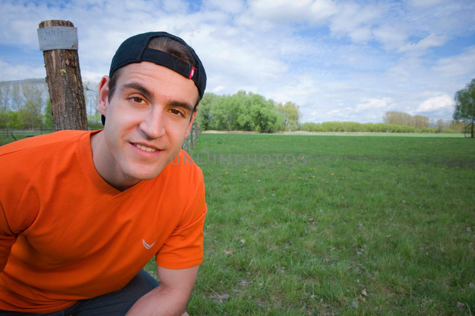 wide-angled near-torso view of a young man in an orange shiver and rear-turned umbrella cap in front of green meadow looking friendly into the camera