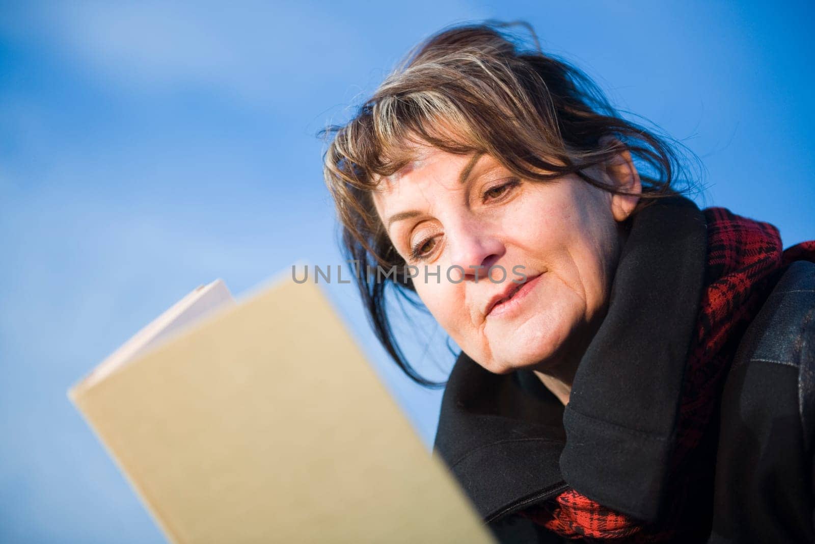 slanted head-and-shoulder view of a mature woman reading in front of blue sky in a book