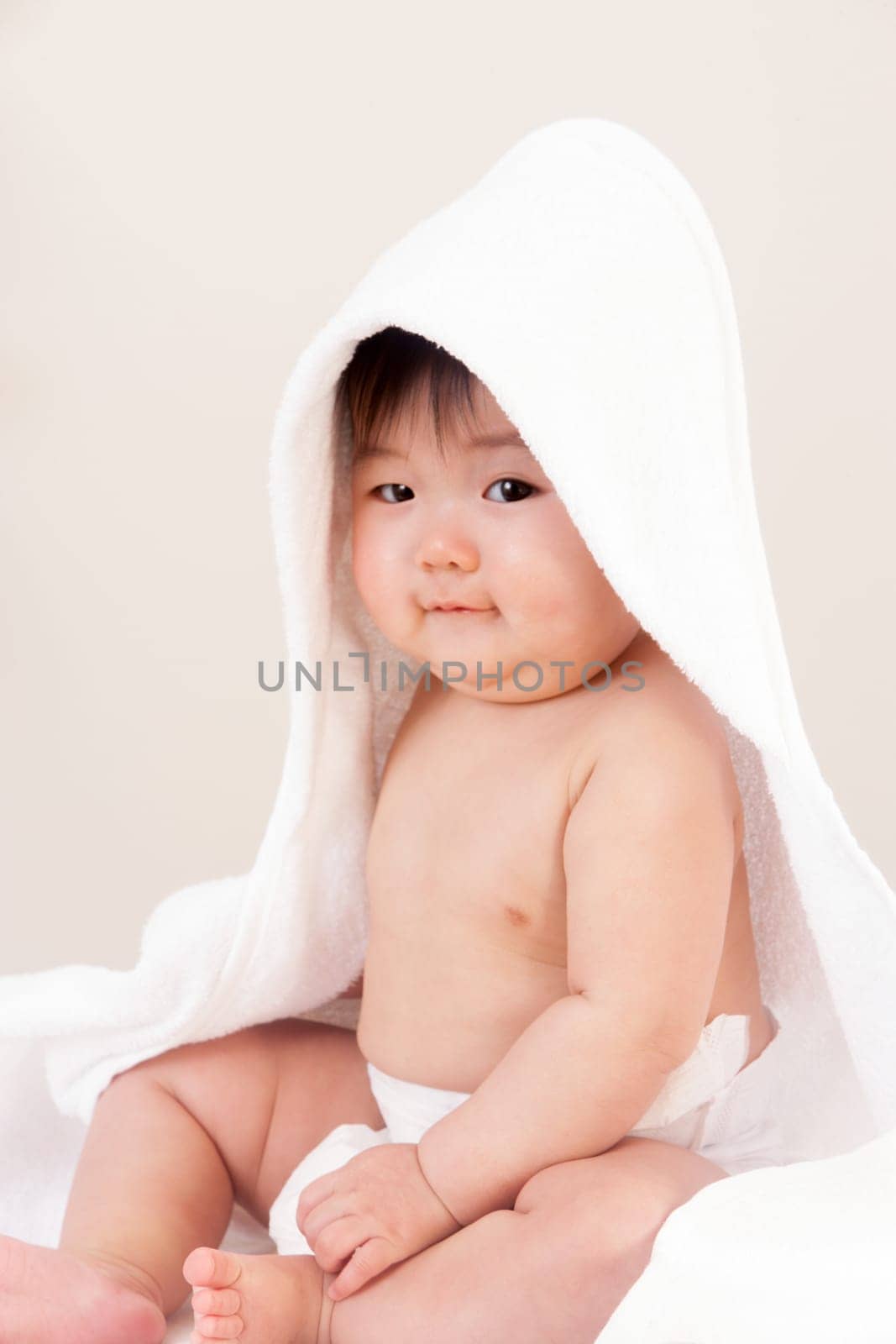 Cute Asian baby sitting with a towel over his head. He is wearing a nappy and is looking at the camera.