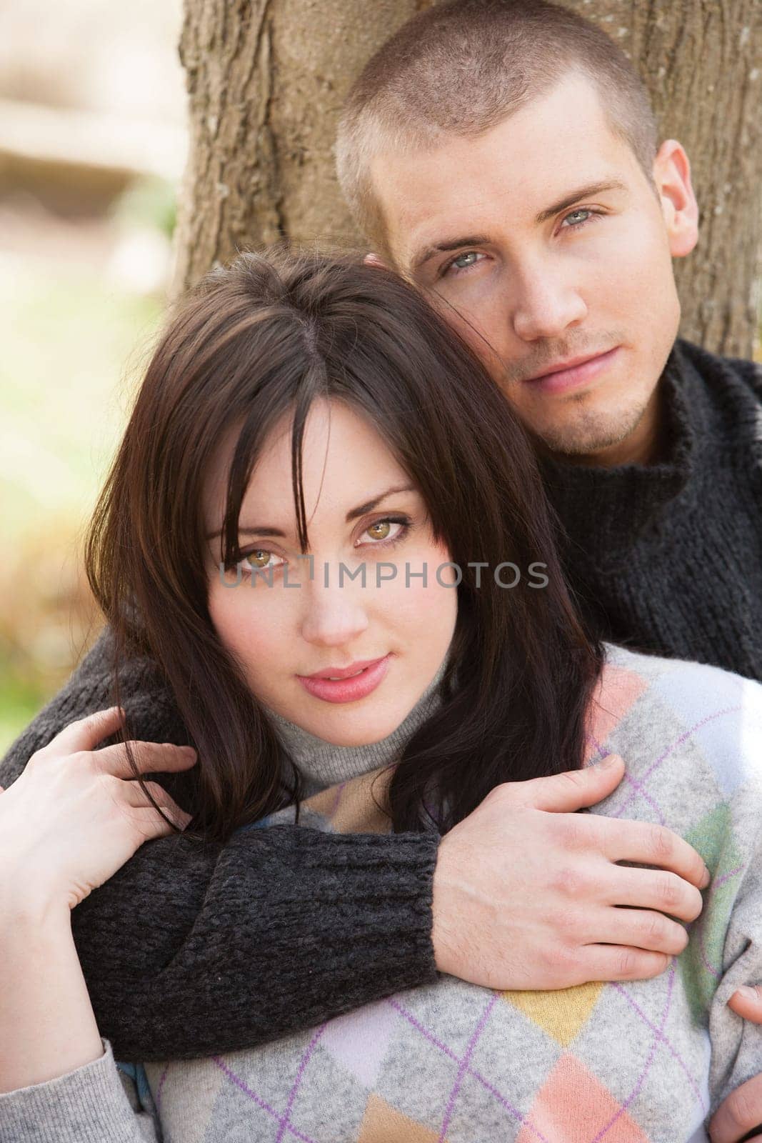 Attractive young couple cuddling out doors. They are both looking at the camera