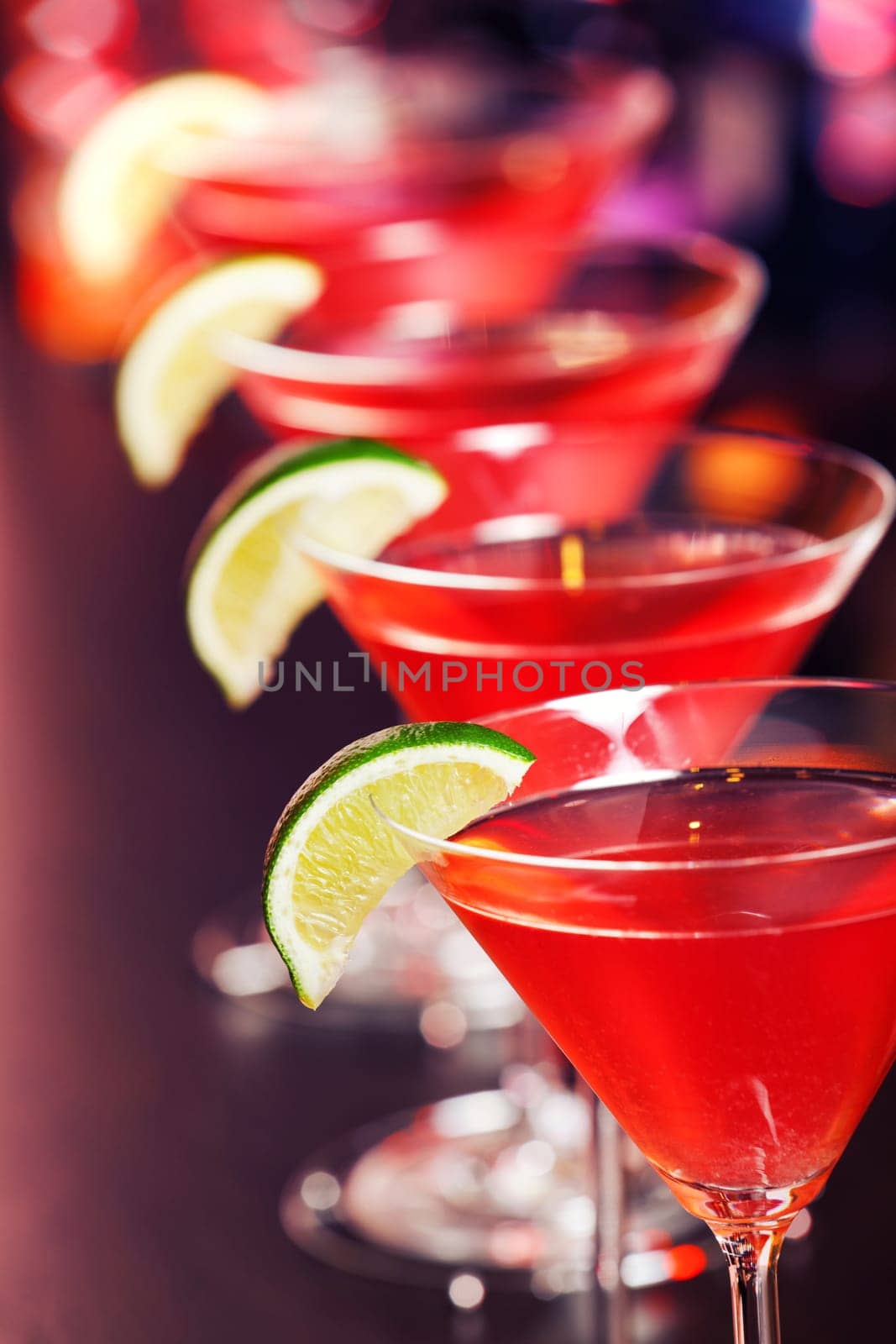 A cosmopolitan, or informally cosmo, is a cocktail made with vodka, triple sec, cranberry juice, and freshly squeezed lime juice or sweetened lime juice. On the photo - four cosmopolitan cocktails in front, blurred bottles of alcoholic drinks in the background.