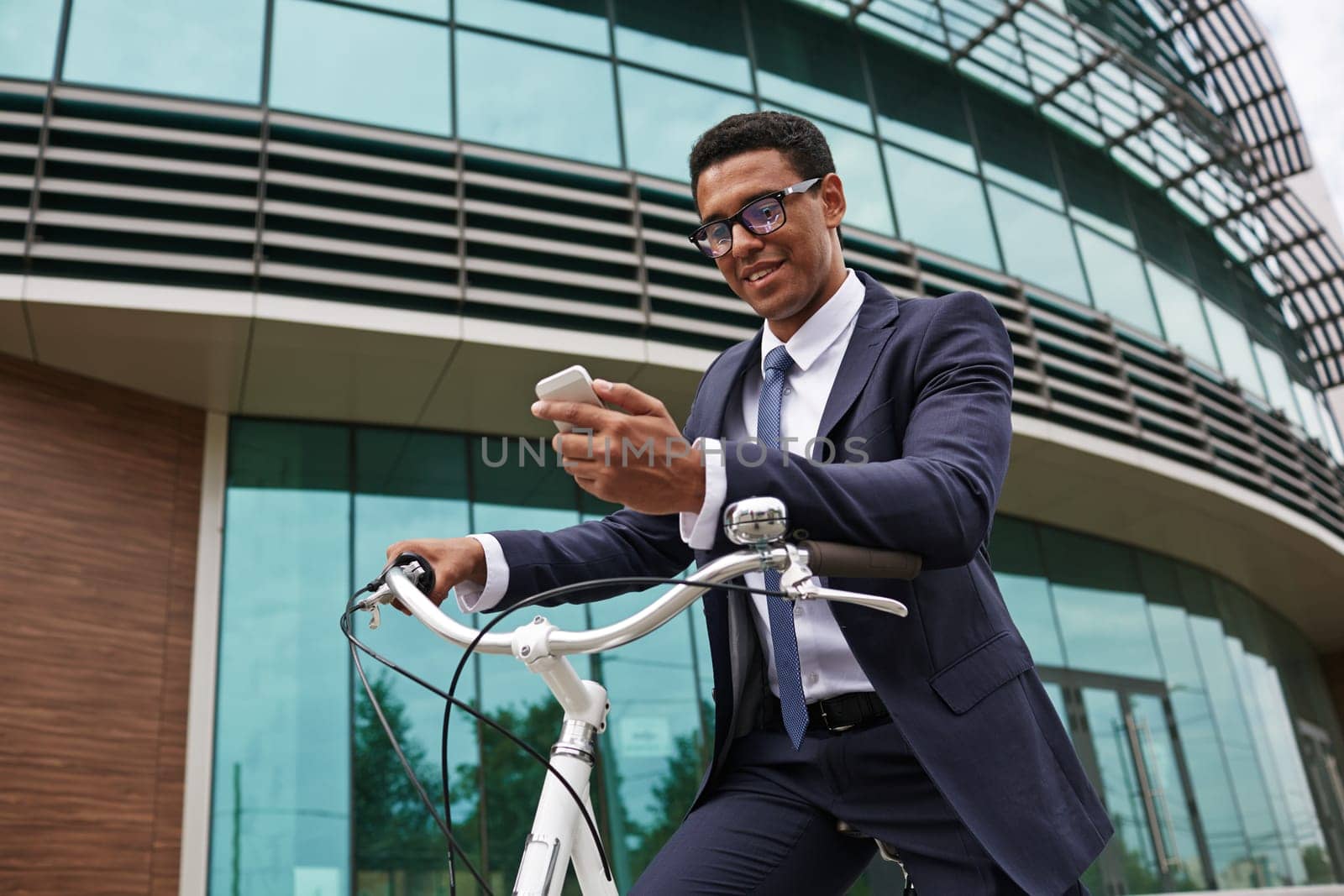 Young businessman sitting on bicycle and reading sms by modern building