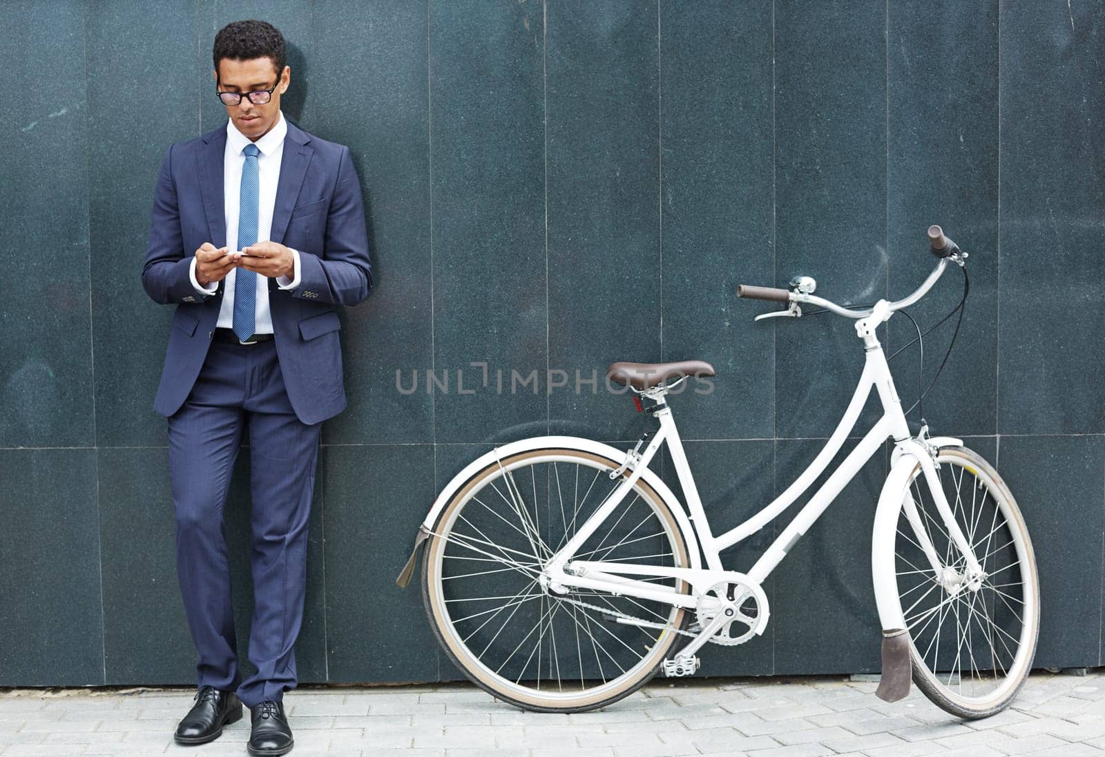 Handsome employee with cellphone standing by wall of modern building with bicycle near by