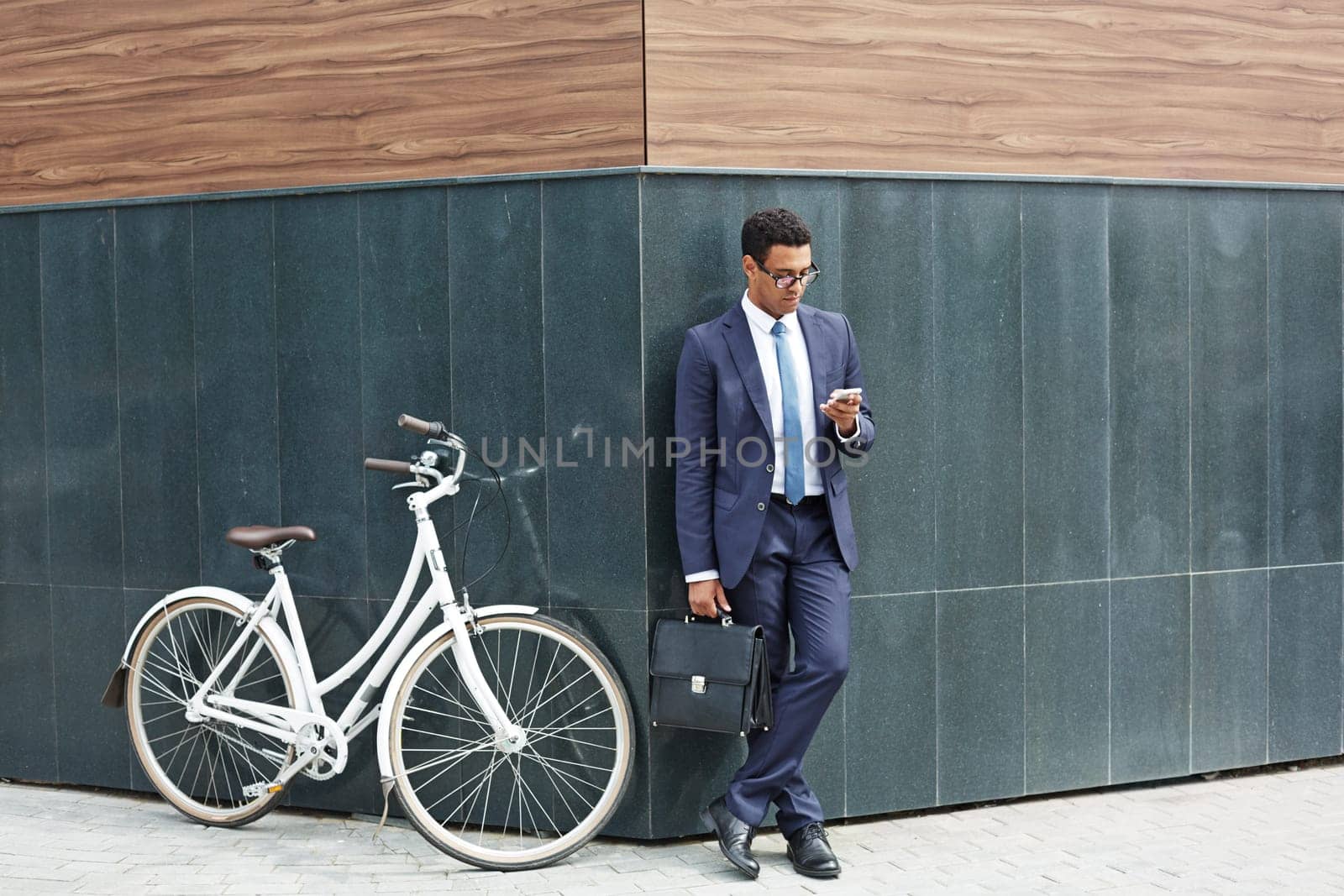 Elegant employee with briefcase using cellphone while standing by corner of modern building with bicycle near by