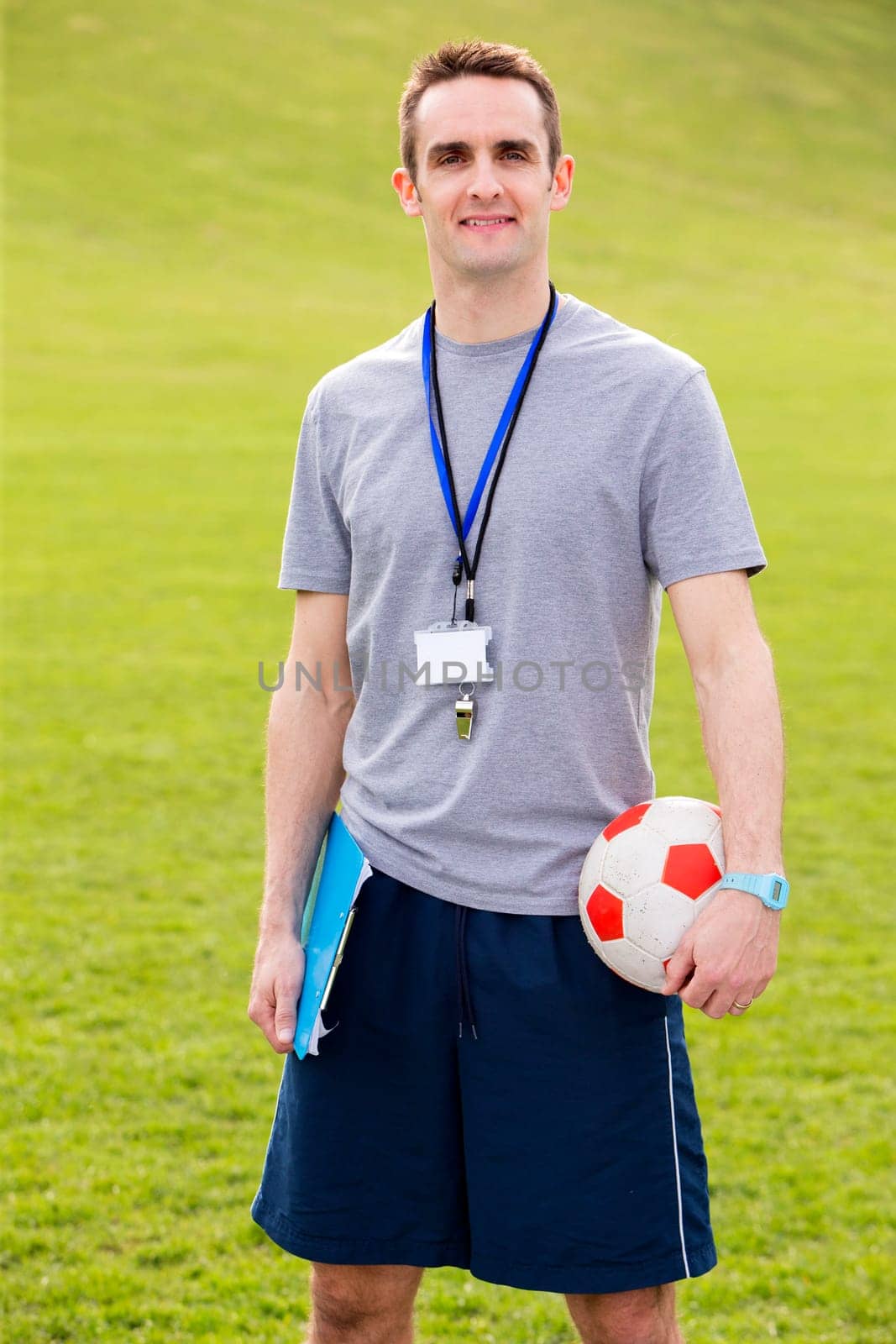A male sports teacher stands outdoors in a field, he is holding a clipboard and football