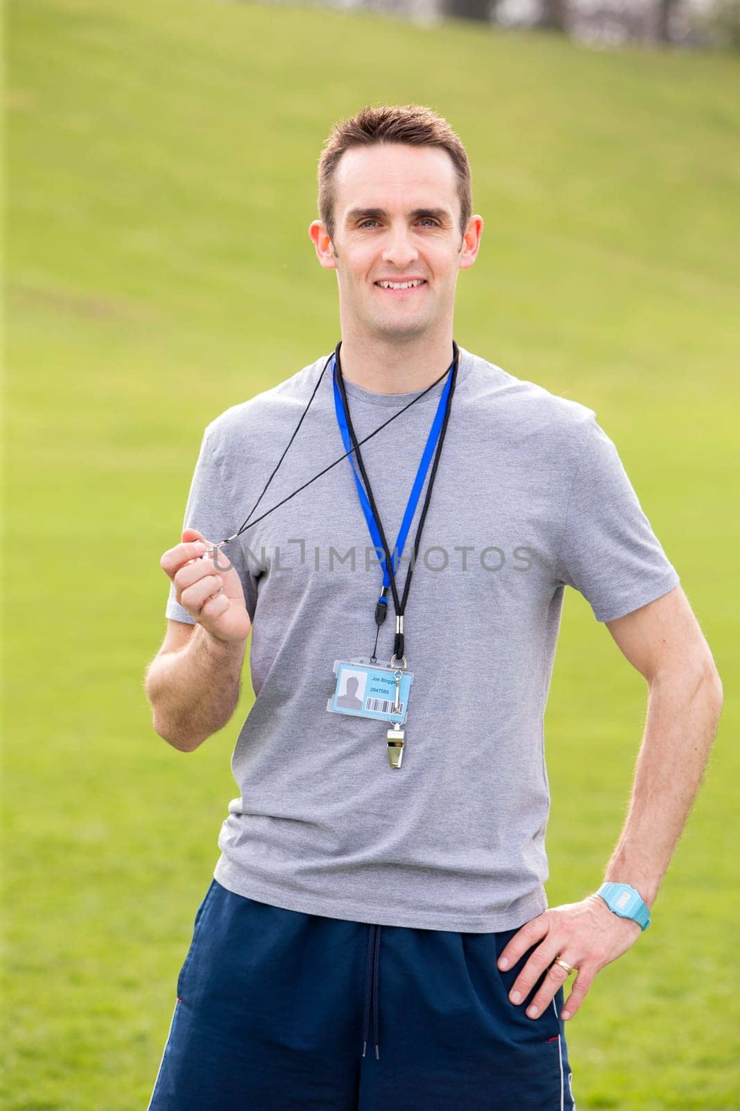 A male sports teacher stands outdoors on a field, he is holding a whistle.