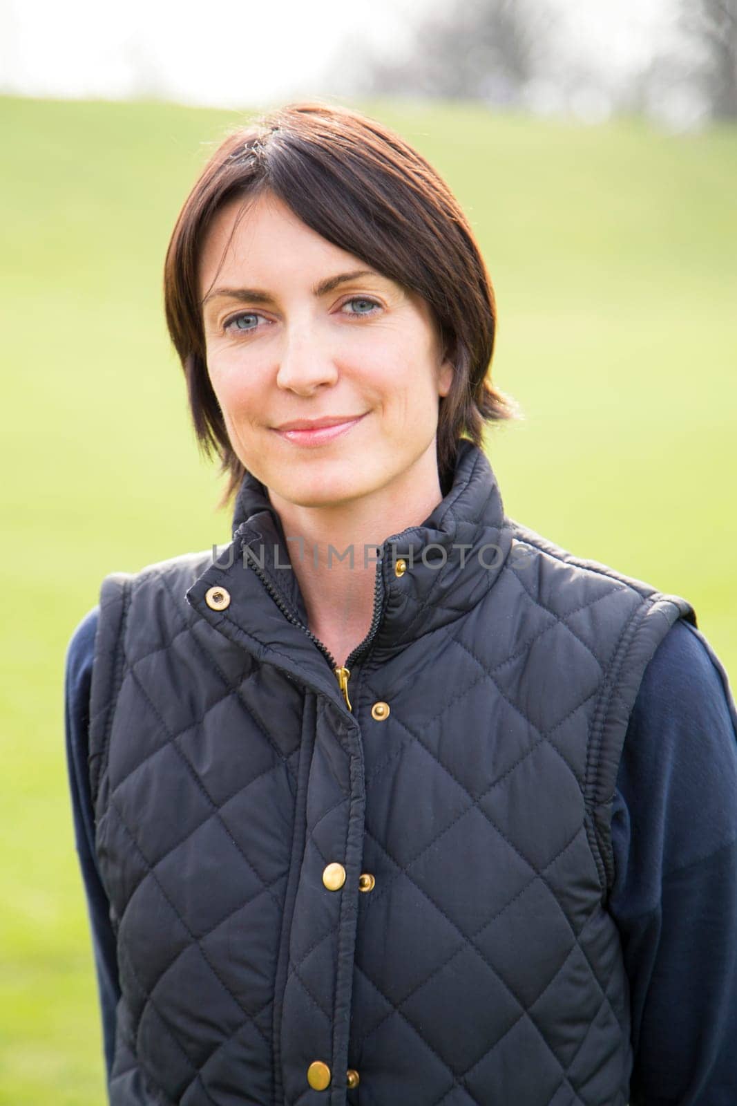 Head and shoulder portrait of a woman looking at the camera, she is outdoors. A grassy area can be seen behind her, She smiles.