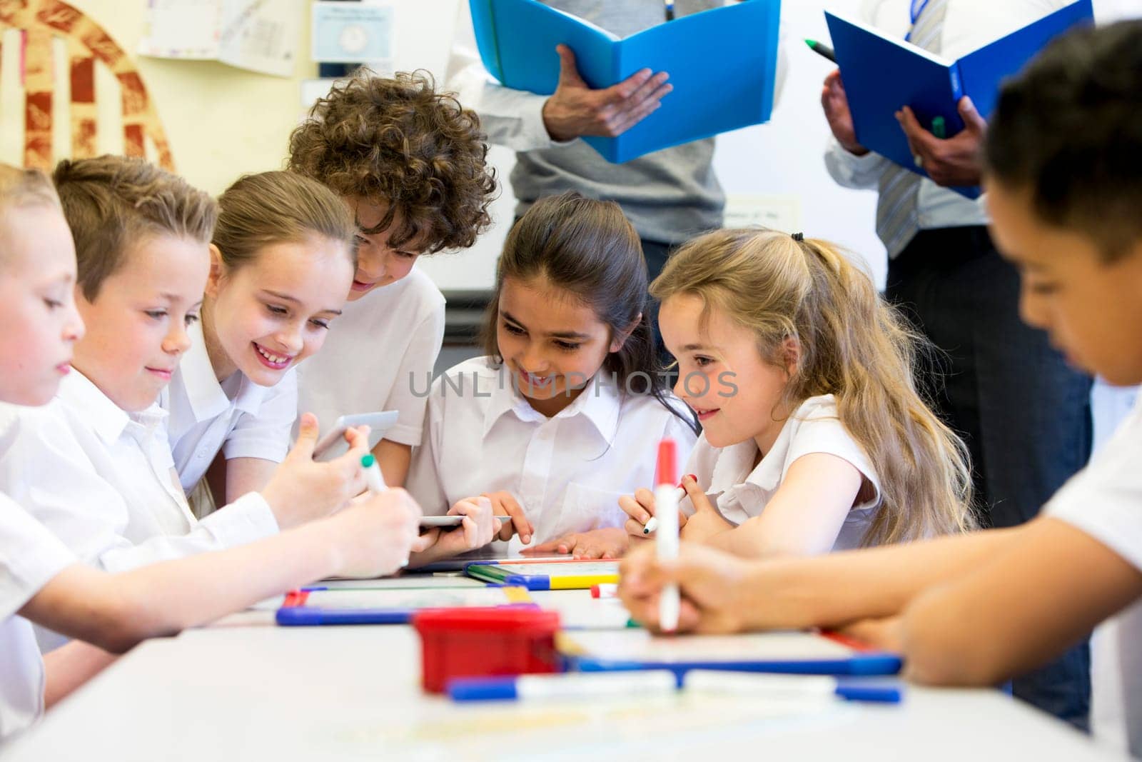 A group of school children can be seen working on digital tablets and whiteboards, they are all working happily. Two unrecognisable teachers can be seen in the background.
