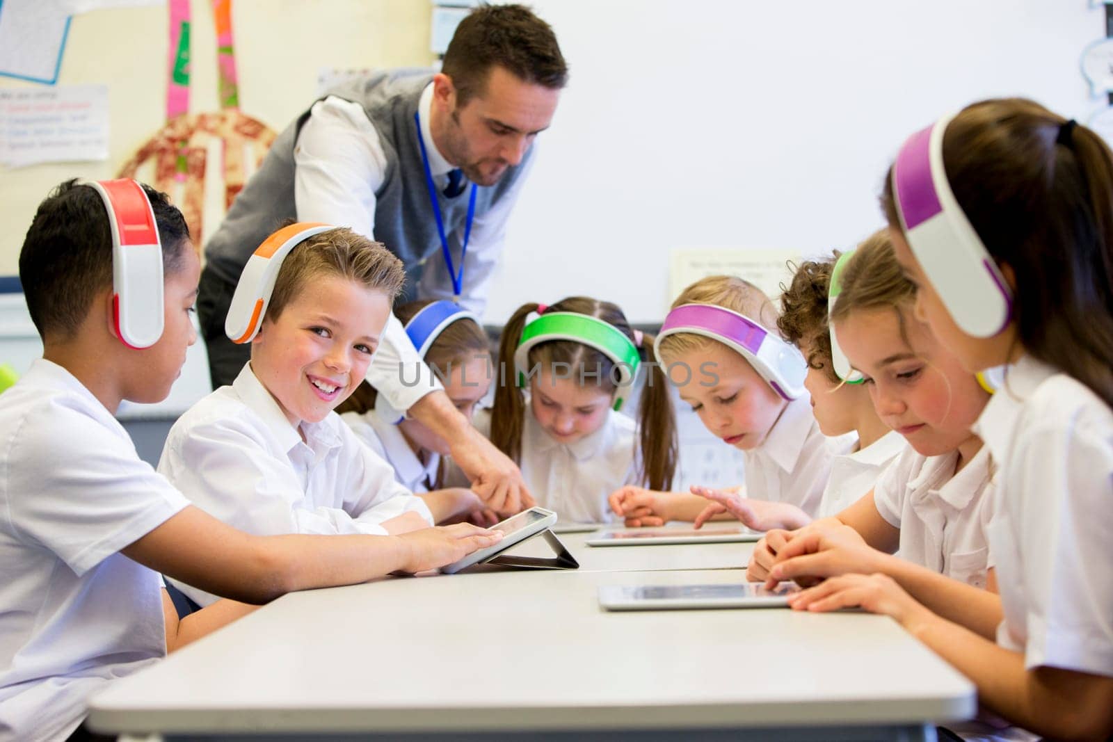 Group of children wearing colourful wireless headsets while working on digital tablets, the teacher can be seen supervising the students in the classroom