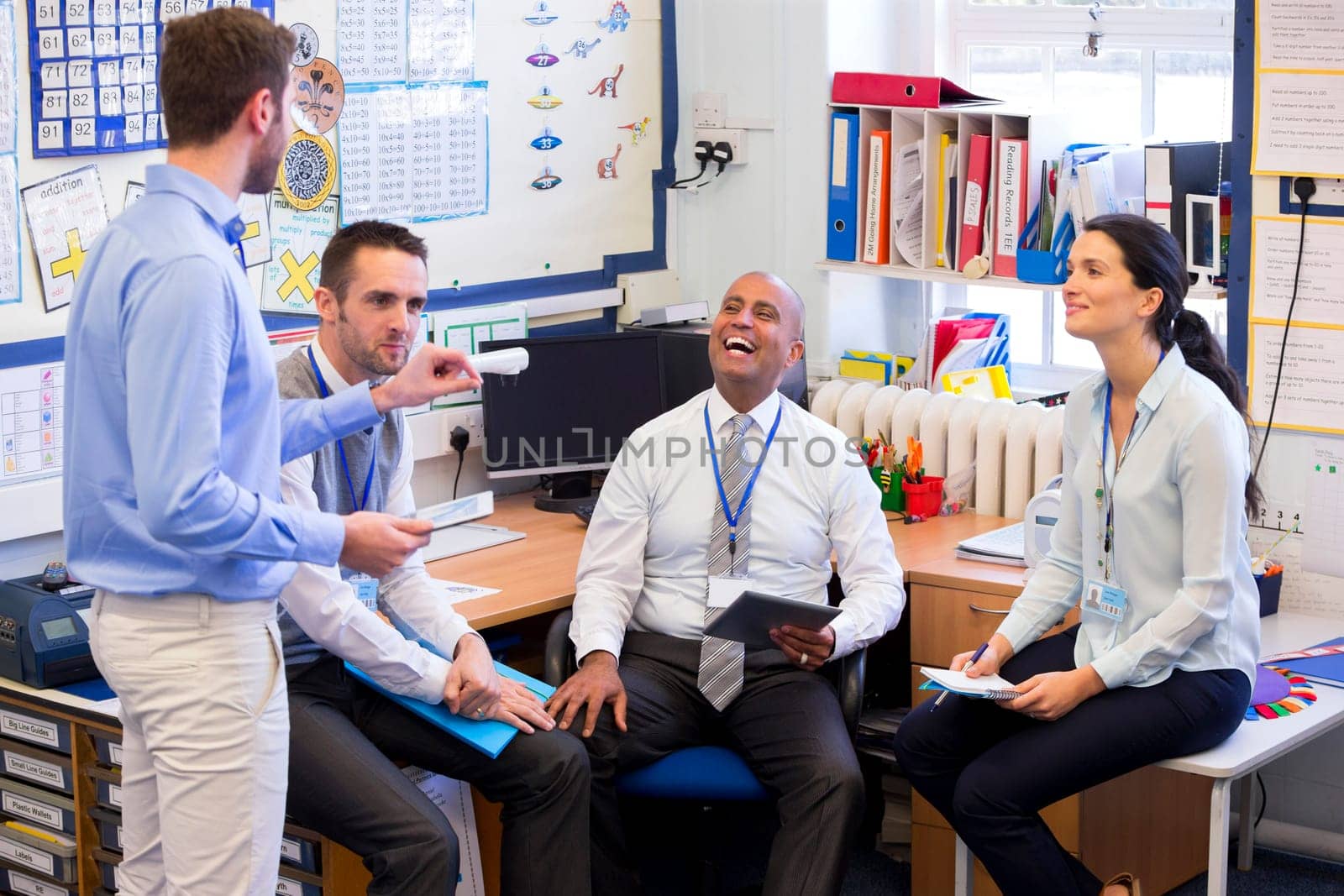 School teachers gather in a small school office for a chat. They look happy. A woman and three men group together.