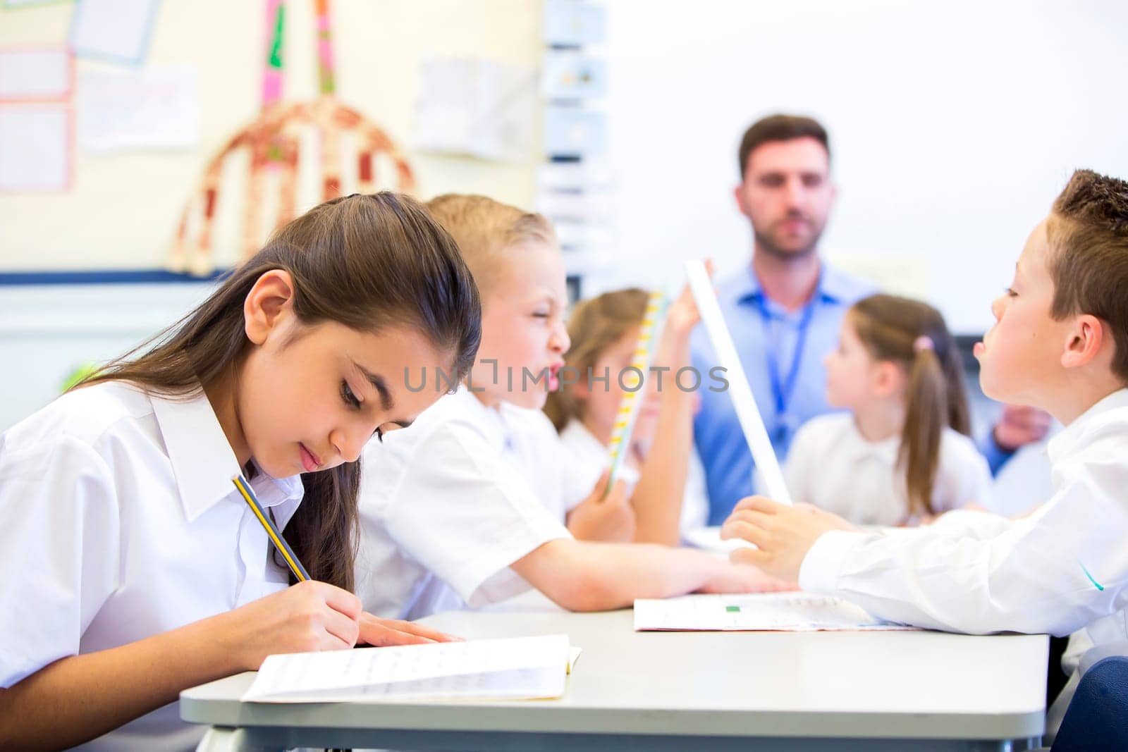 Schoolgirl sitting at a desk with other classmates while working. Her head is down while she concentrates. The teacher can be seen in the background.