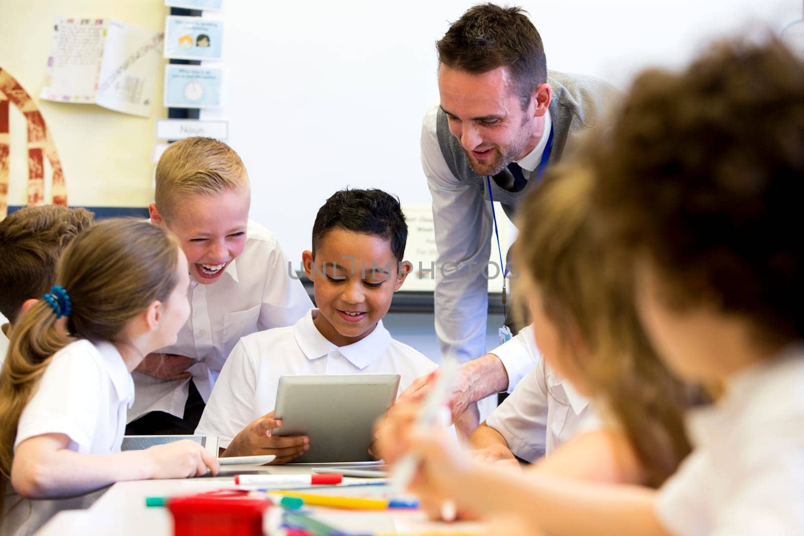 A male teacher sits supervising a group of children who are working on whiteboards and digital tablets.
