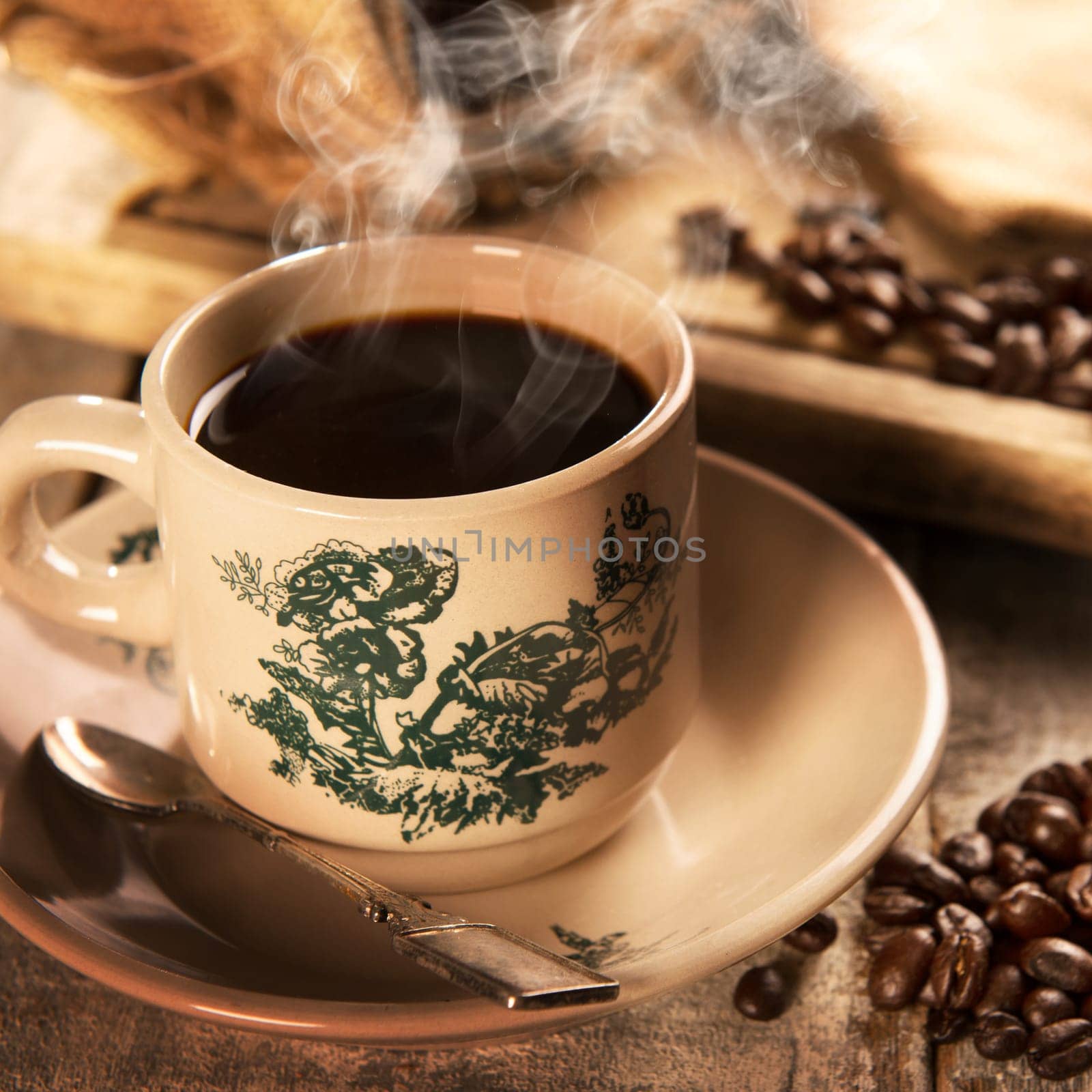 Steaming traditional Hainan style dark coffee in vintage mug and saucer with coffee beans. Fractal on the cup is generic print. Soft focus setting with dramatic ambient light on dark wooden background.