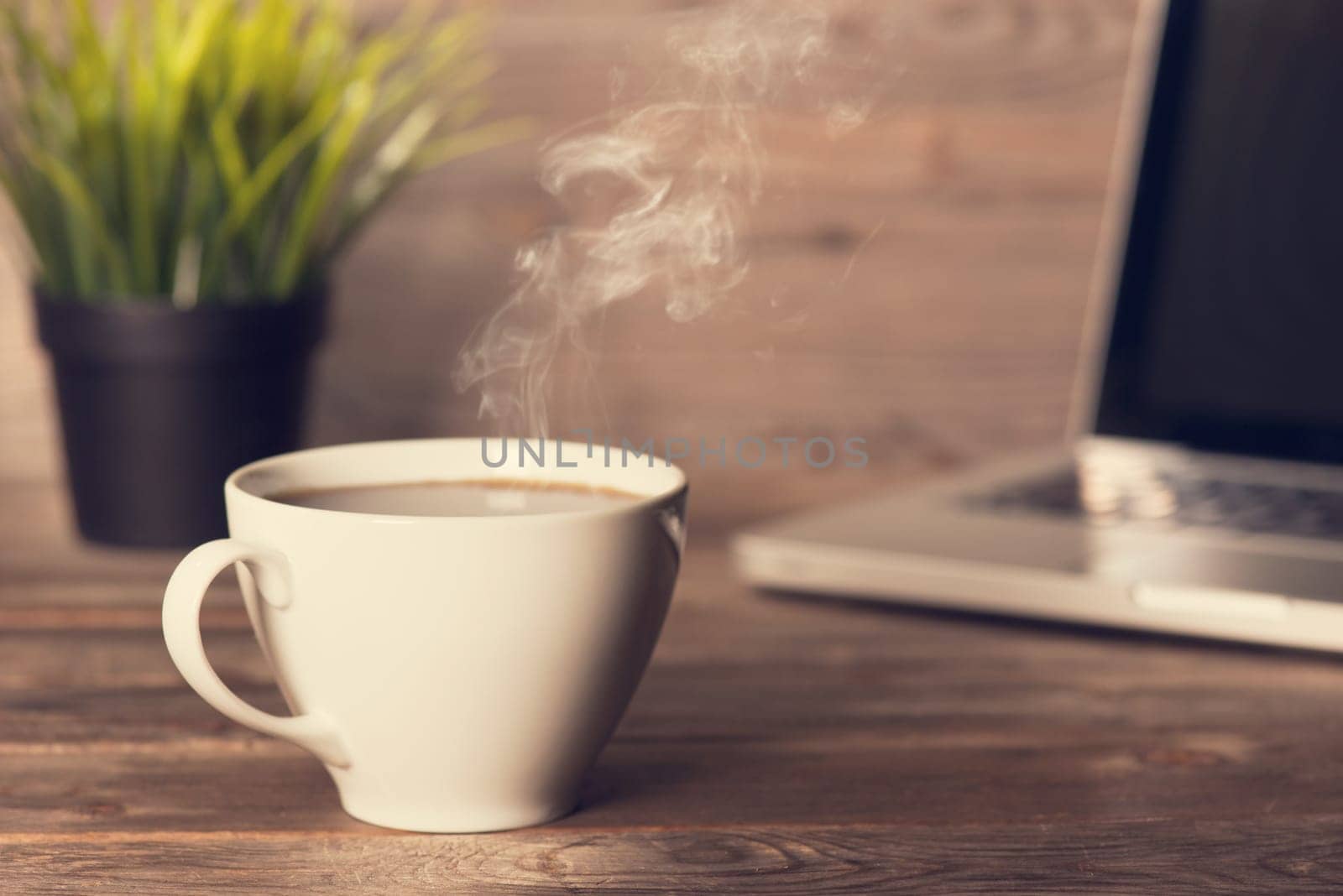 Morning caffeine concept. Wooden business desk with laptop, cup of hot coffee and pot plant, in dramatic light vintage toned.