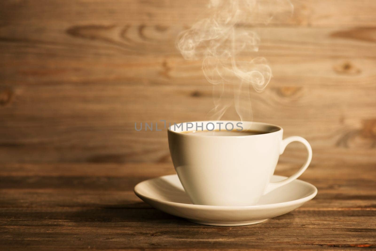 Steaming coffee in white mug and saucer in soft focus setting with dramatic ambient light, over dark wooden background.
