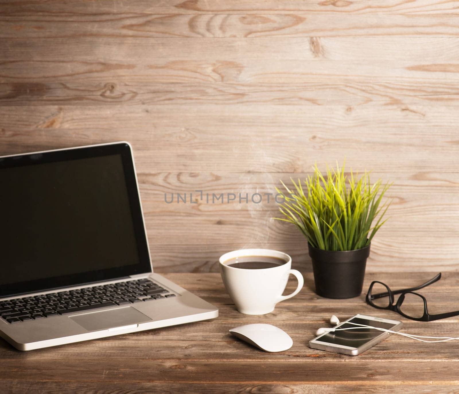Wooden office interior, table with laptop, cup of hot coffee, mouse, glasses, smartphone, earphones and pot plant, in dramatic light vintage toned.