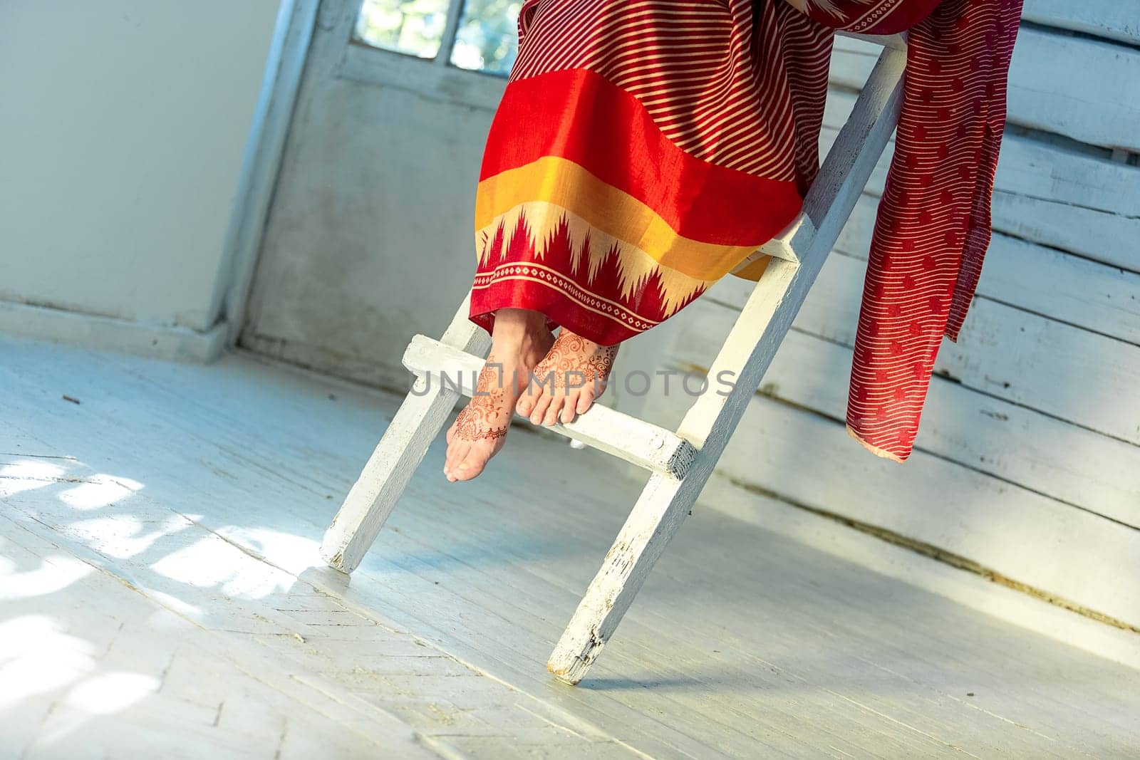 Indian picture on woman feet, mehendi tradition decoration, resistant design by special paint, brown henna. Woman sitting in indian red dress on white wooden staircase background