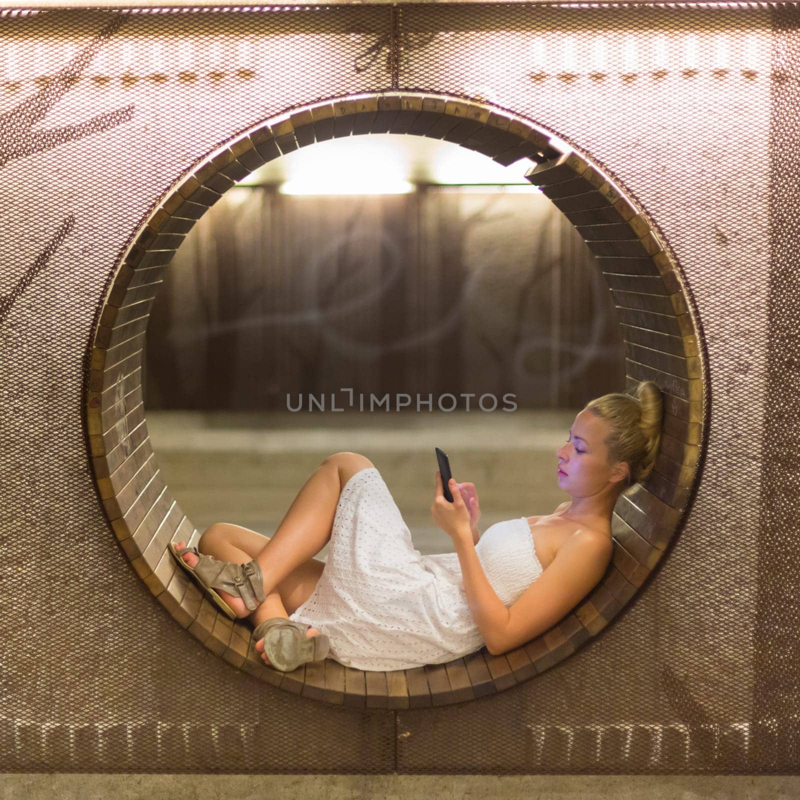 Lady wearing white summer dress playing carelessly with her android smart phone browsing trough social networks relaxing on contemporary circular bench in urban pedestrian underpass.