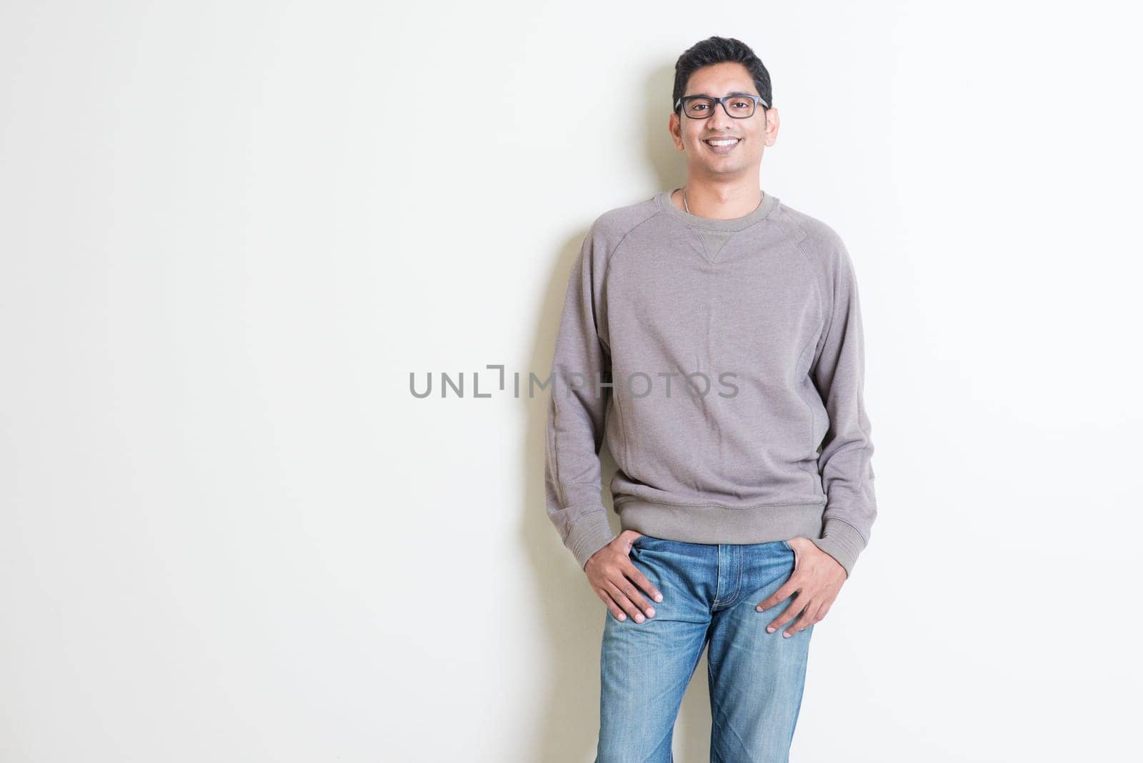 Portrait of handsome casual Indian male smiling, standing on plain background with shadow, copy space at side.