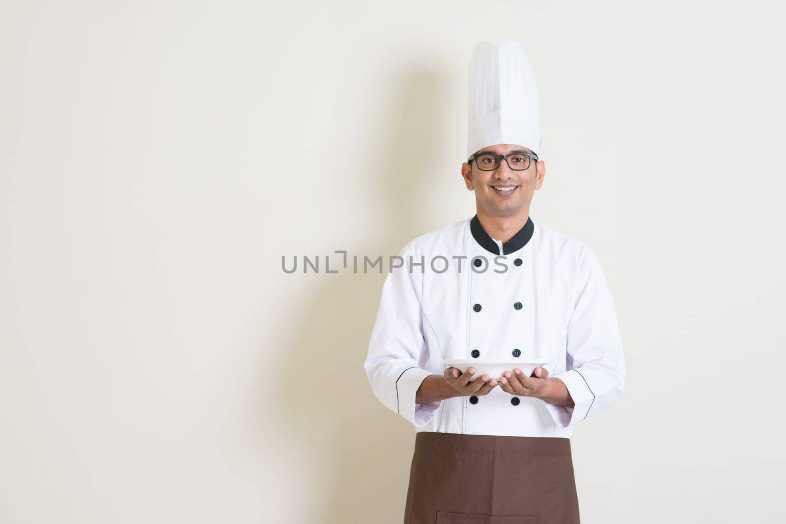 Portrait of handsome Indian male chef in uniform hands holding an empty plate and smiling, standing on plain background with shadow, copy space.
