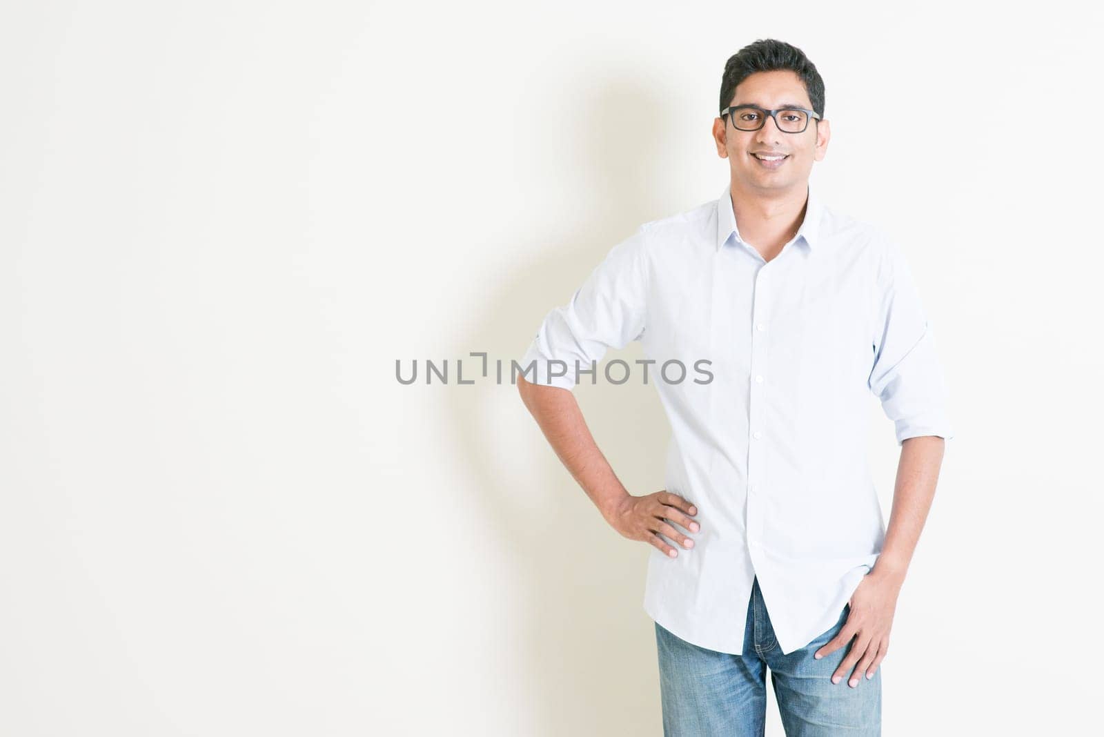 Portrait of handsome casual business Indian male smiling, standing on plain background with shadow, copy space at side.