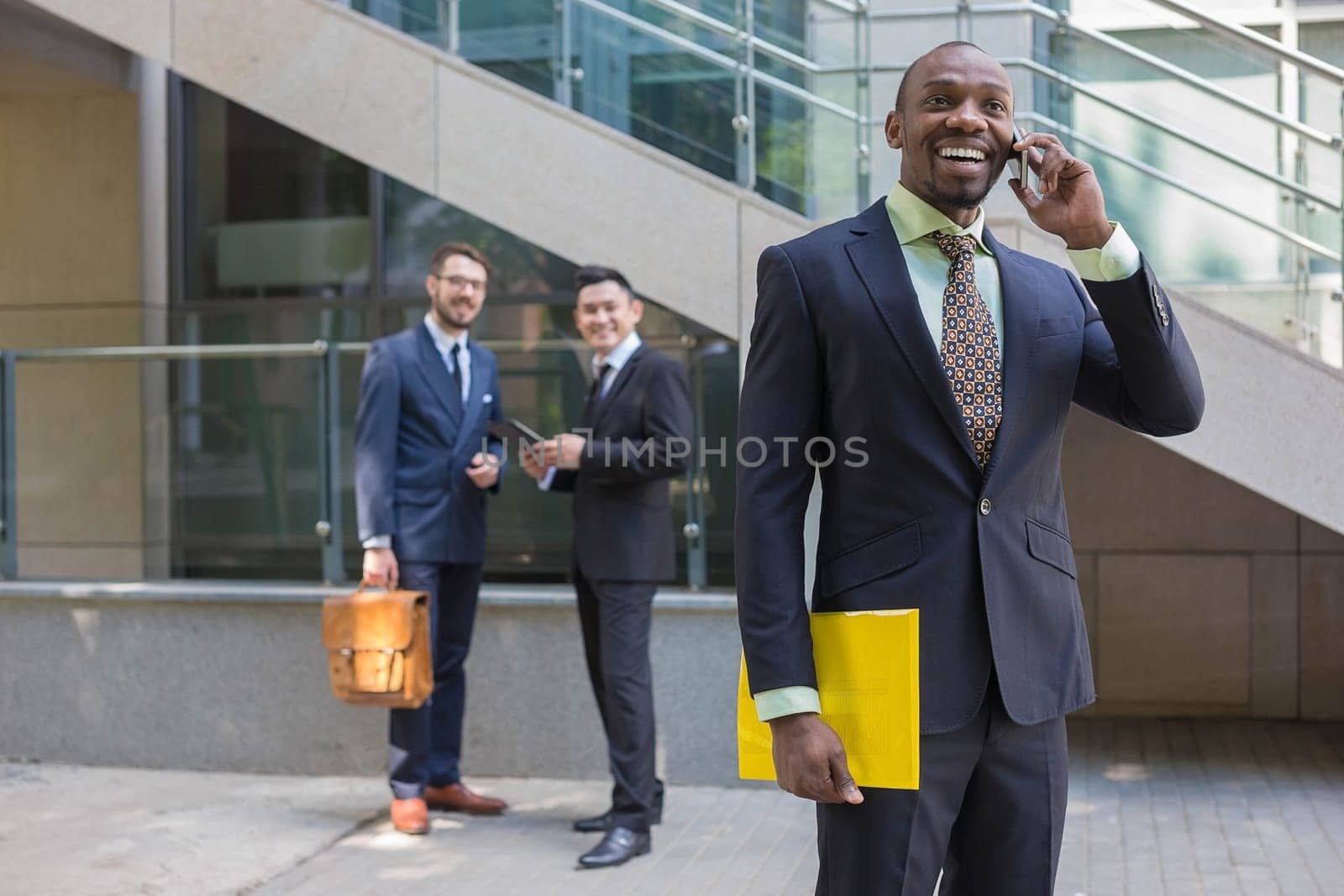 Portrait of multi ethnic business team. Three happy smiling men standing against the backdrop of the city.  The foreground of a African-American man talking on the phone