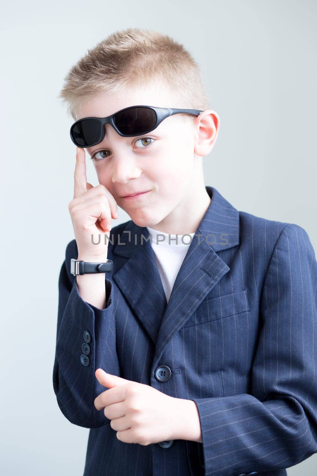 Young boy dressed up and posing like a spy. He is wearing a suit with sunglasses on his head and a formal watch. He is miming a gun with his hands.