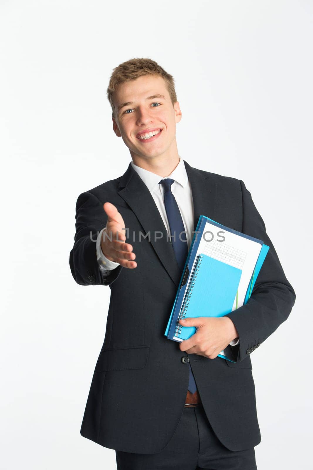 Young businessman holding his hand out to offer a handshake. He is dressed in a suit and is holding folders whilst standing against a plain background