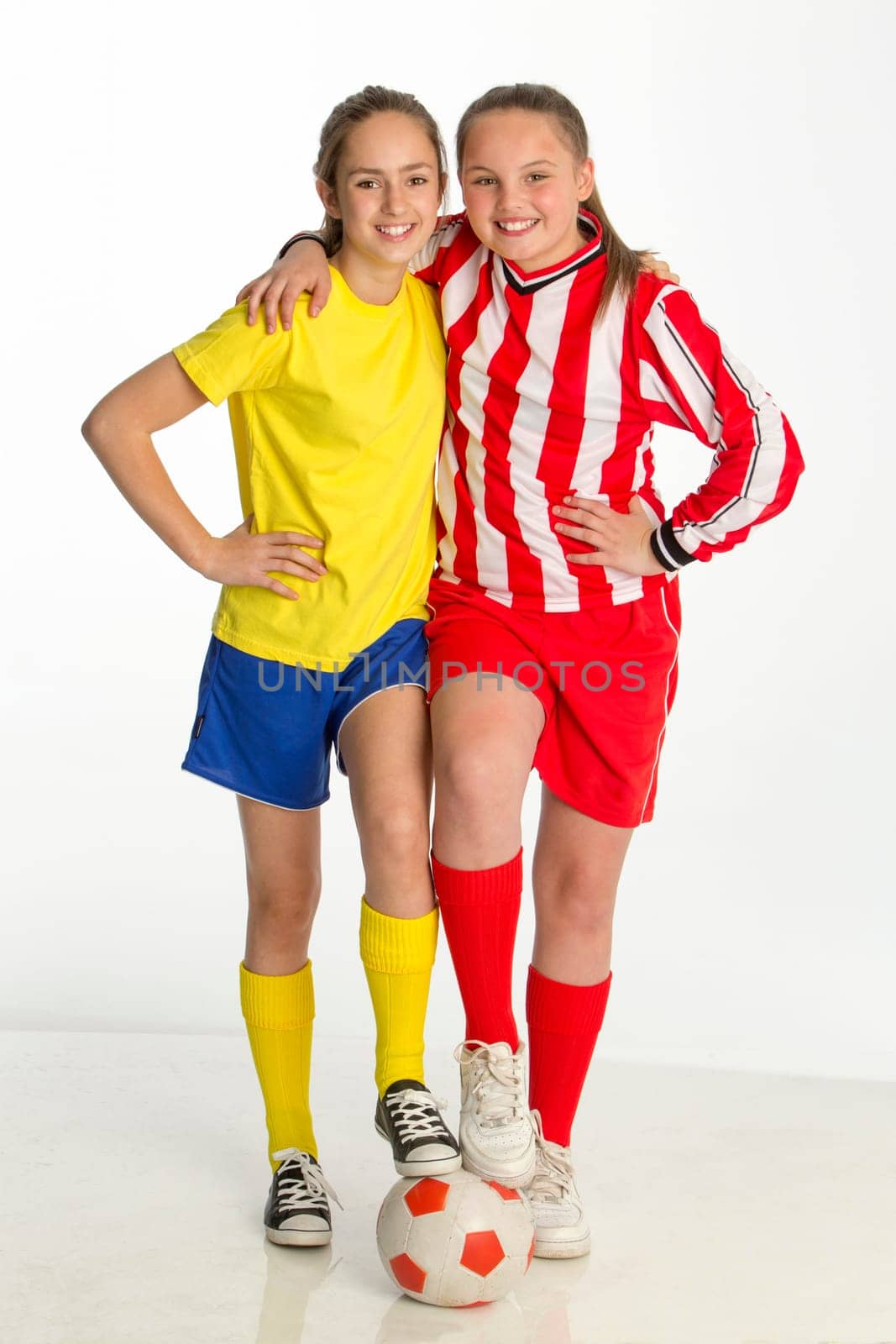 Team captains of girls football match posing with a ball against a white background. They both have one foot on the ball and one arm round each other whilst smiling for the camera.