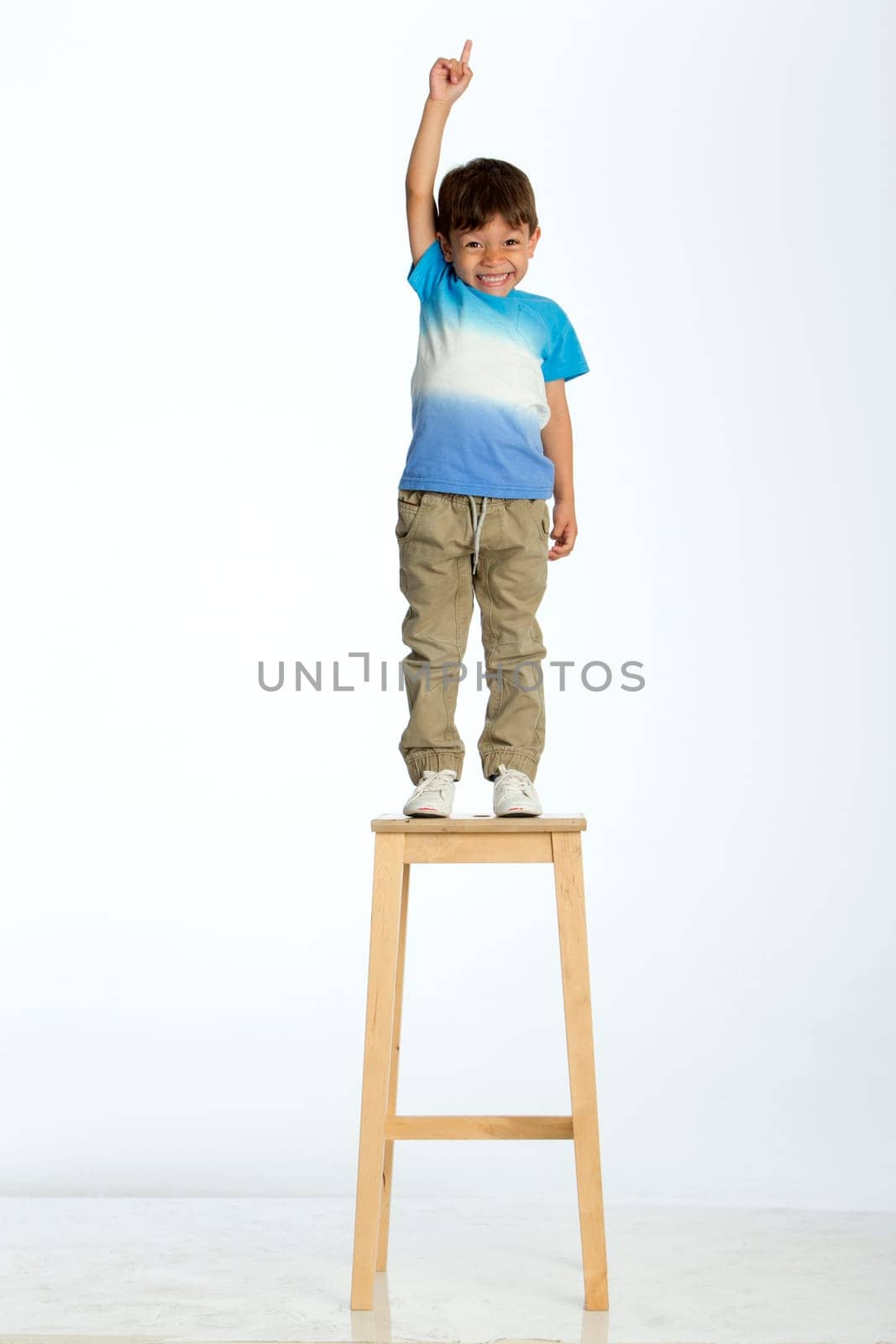 Little boy standing on a high stool, pointing up to the ceiling. He is against a white background.