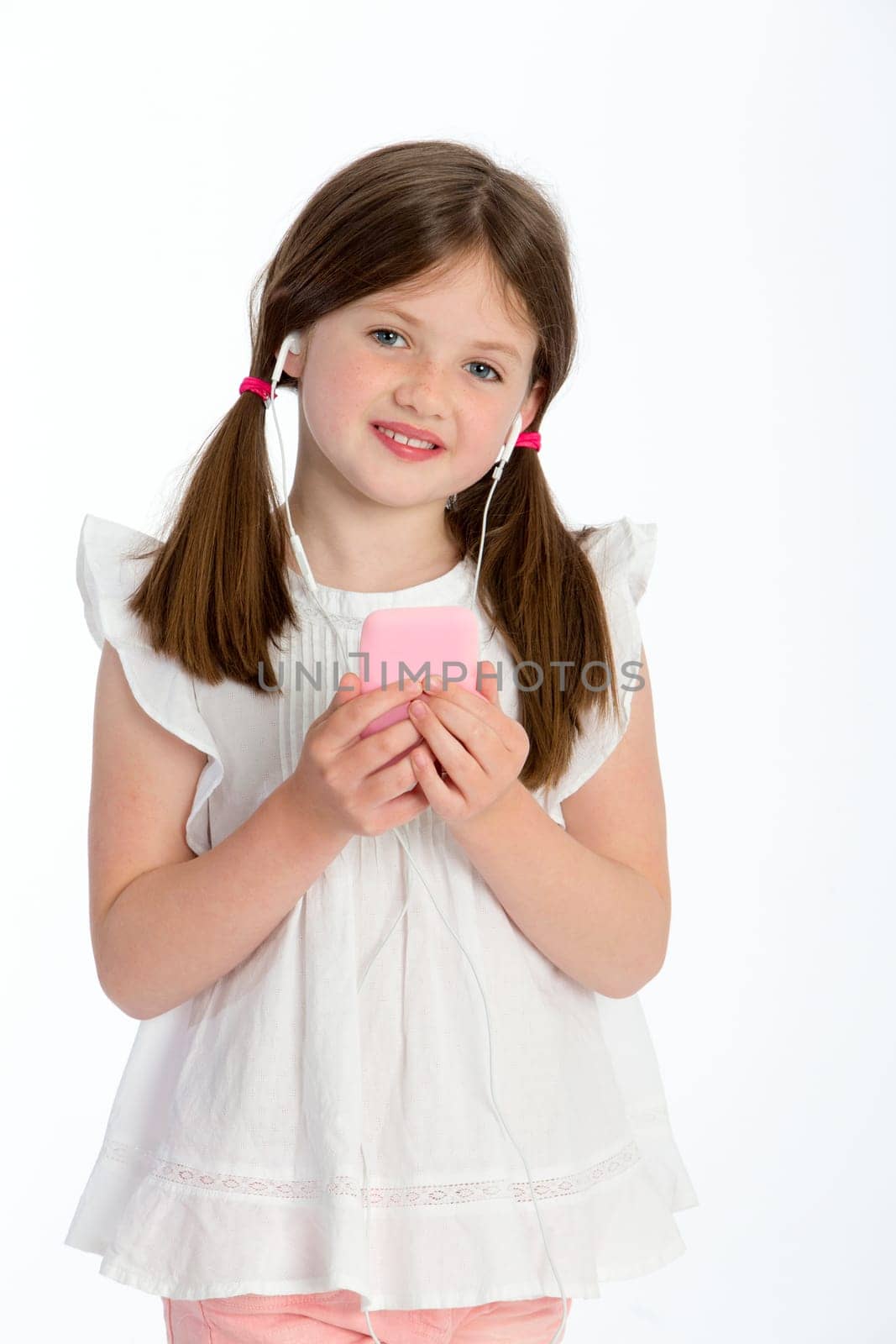 Little girl listening to music through headphones on a smartphone. She is smiling at the camera and standing against a white background.