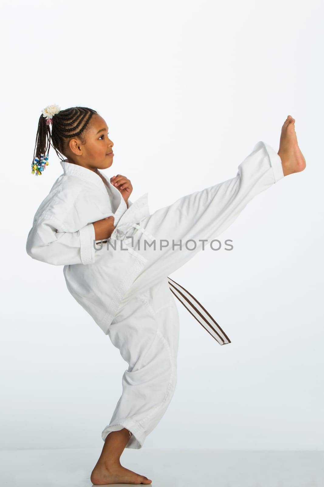 Young mixed race girl posing in a karate kick position. She is wearing a karategi and is standing against a white background.