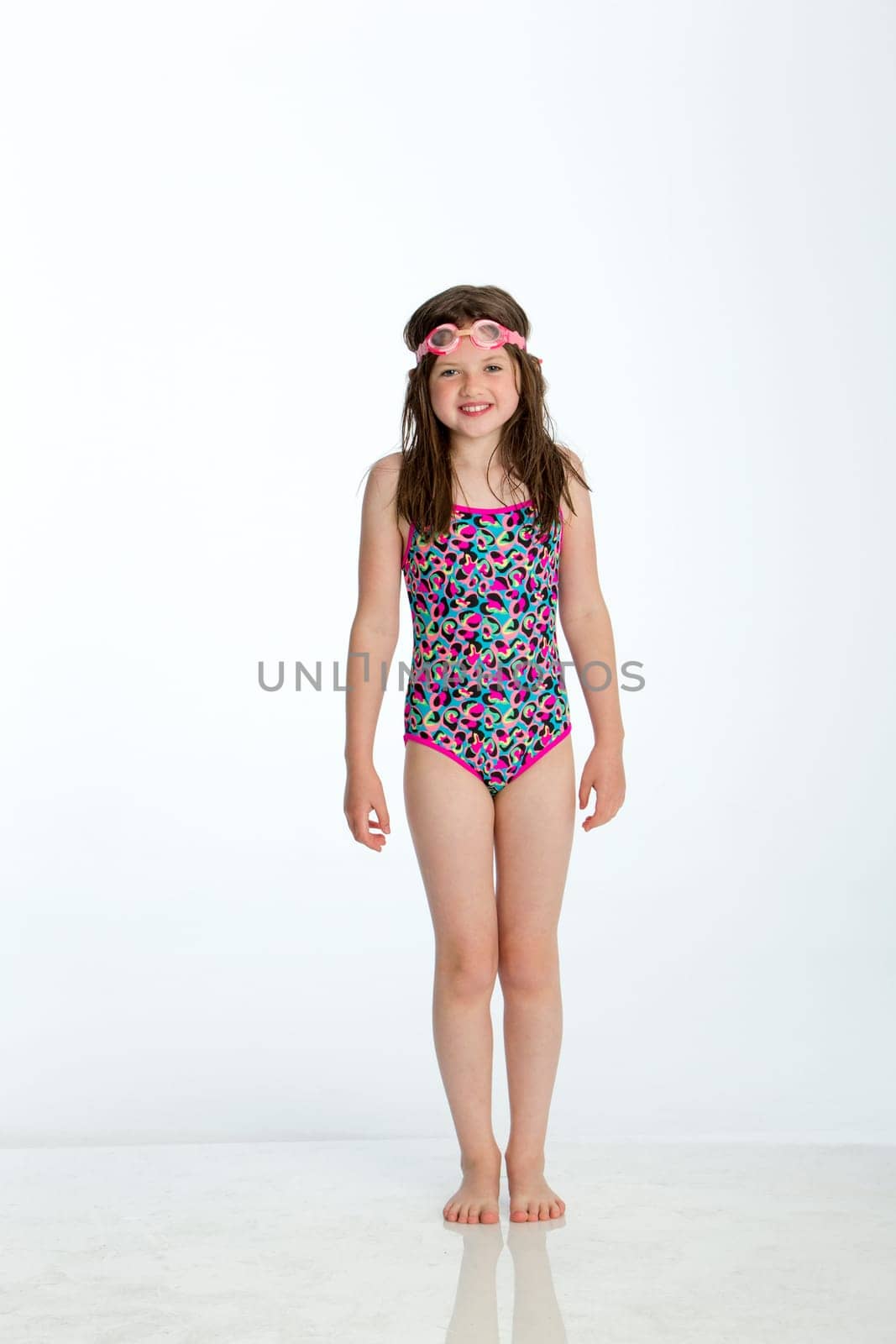 Full length shot of a little girl wearing a swimming costume with goggles on her head. She is smiling at the camera and standing against a white background.
