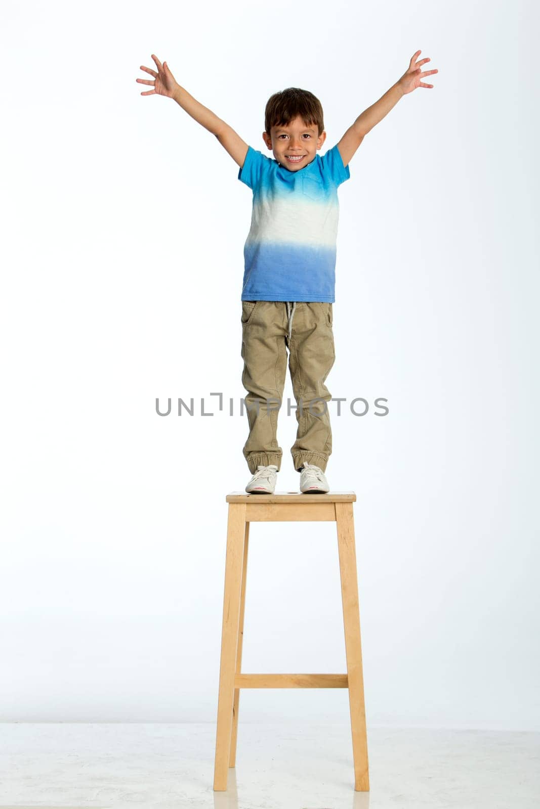 Little boy standing on a high stool with his arms out stretched. He is against a white background.