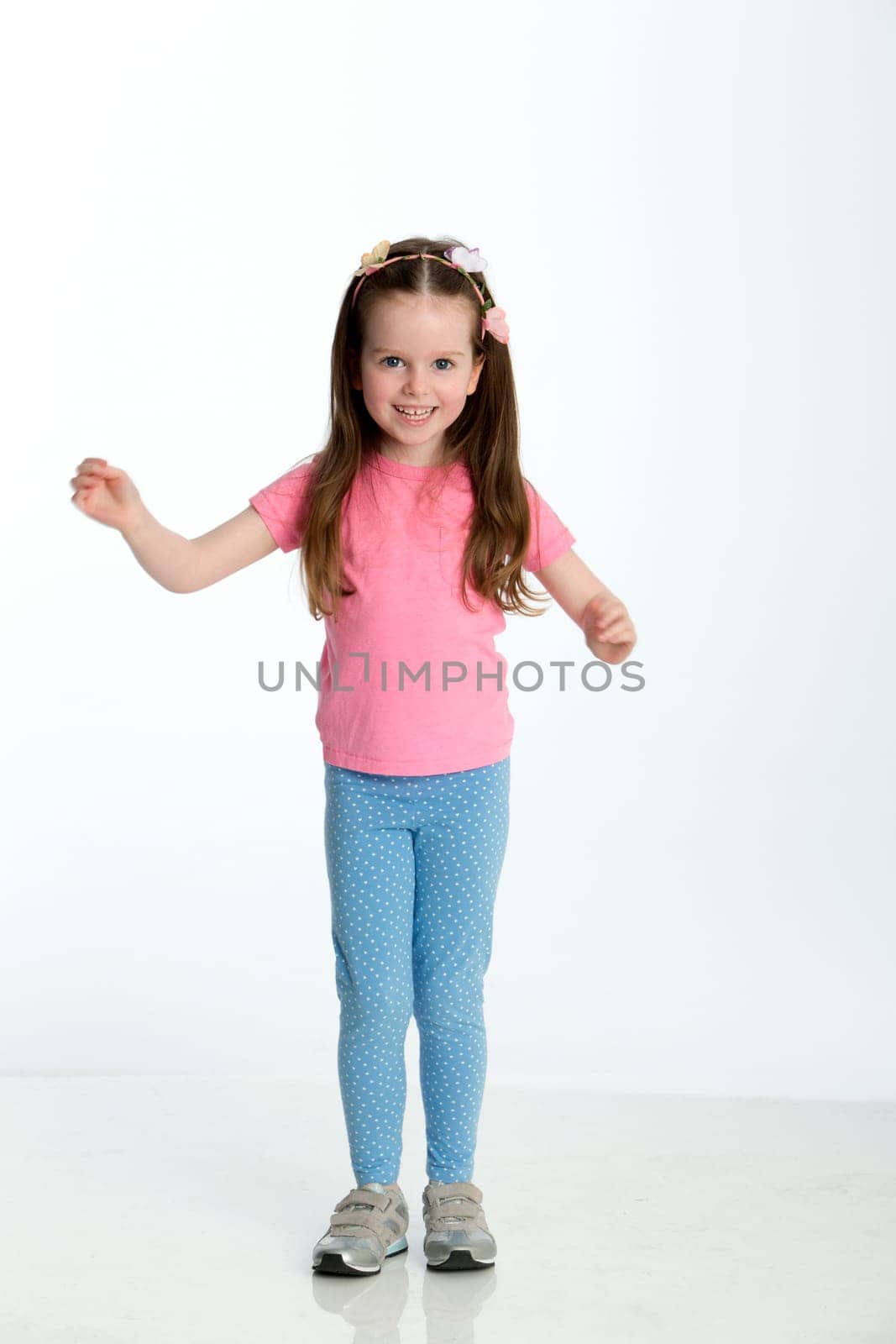 Young girl posing for the camera with her arms out. She is standing against a white background and is smiling at the camera.