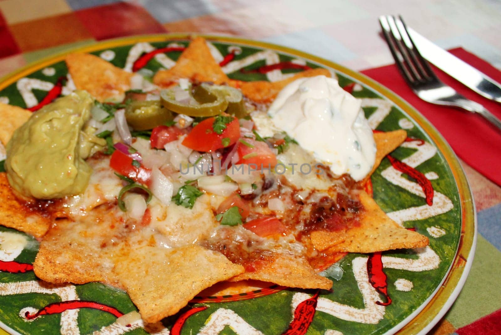 A plate of delicious tortilla nachos with melted cheese sauce, ground beef, jalapeno peppers, onions, tomato, cheese, salsa, and sour cream with guacamole dip