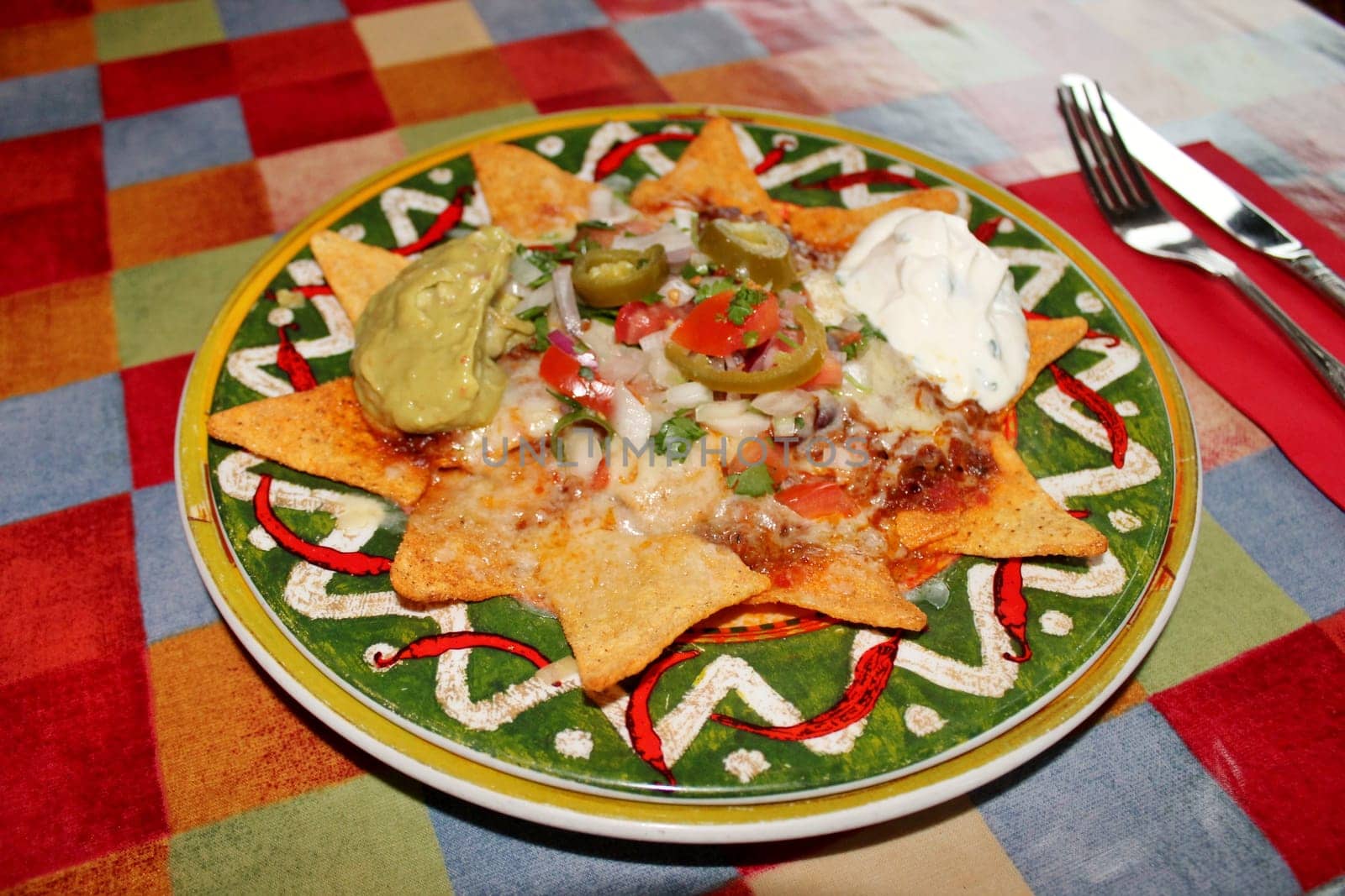 A plate of delicious tortilla nachos with melted cheese sauce, ground beef, jalapeno peppers, onions, tomato, cheese, salsa, and sour cream with guacamole dip