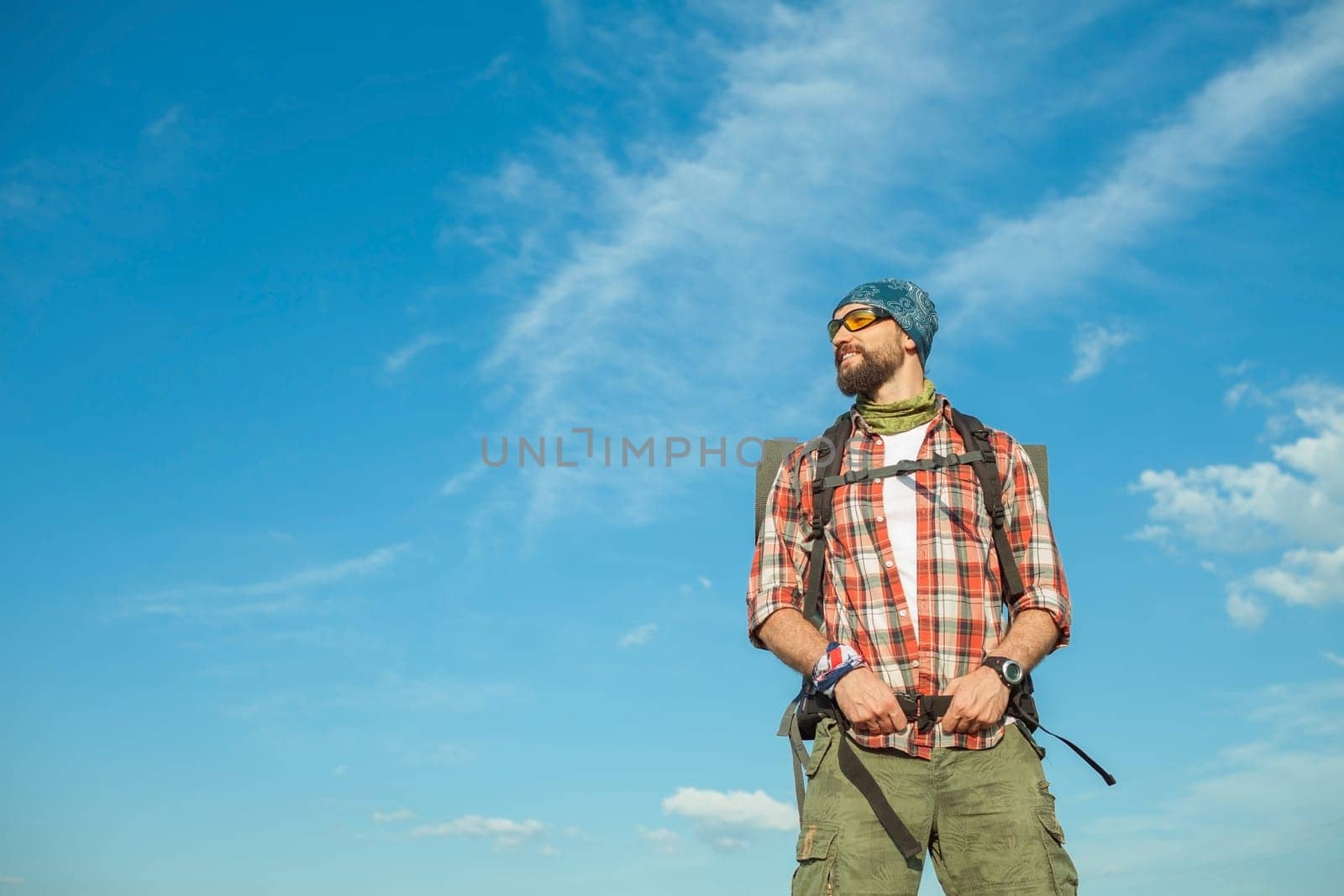 Young smilimg caucasian tourist with backpack  standing on the top of hill against blue sky background. The concept of an invitation to travel