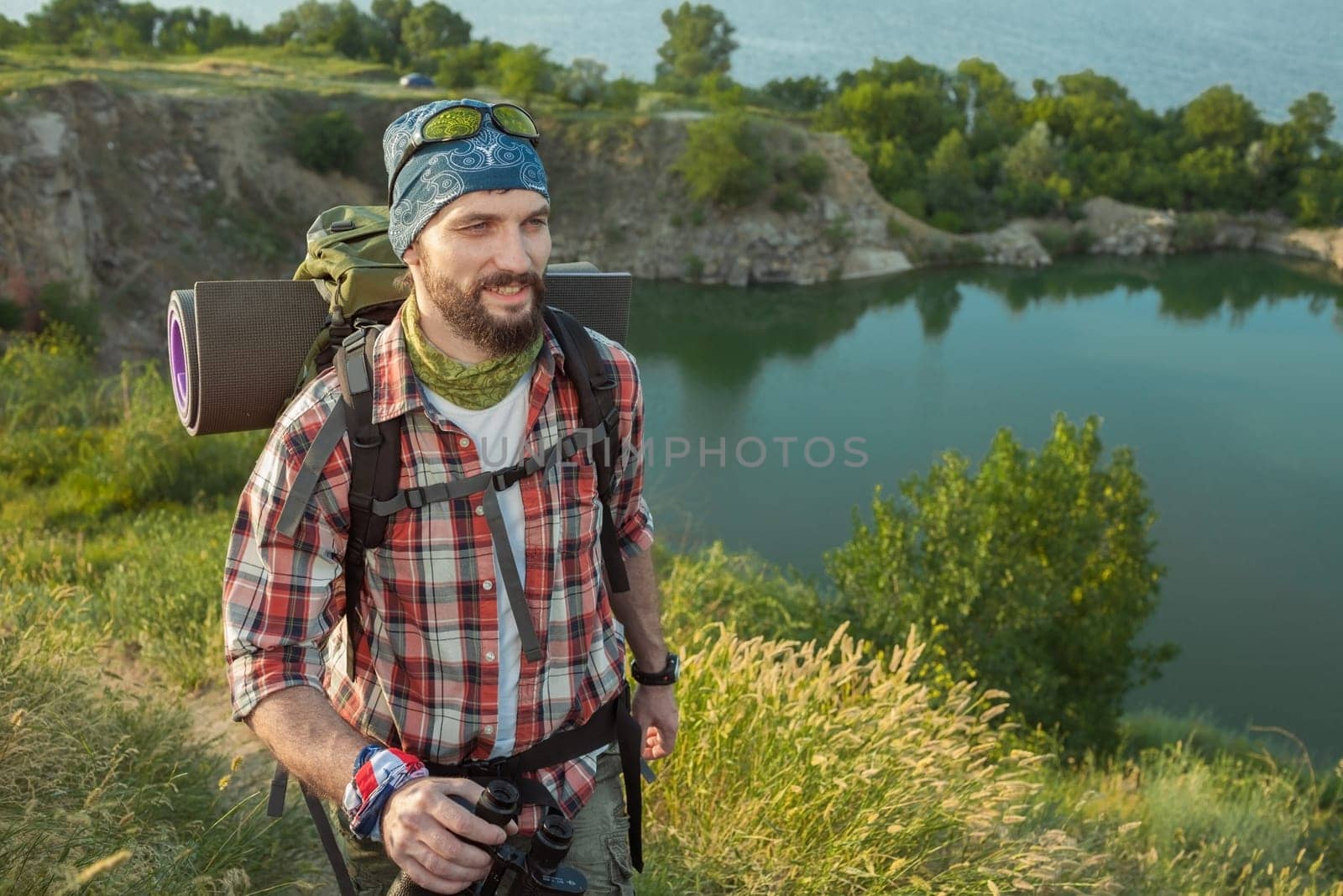 Young smilimg caucasian tourist with backpack walking on the top of hill and looking at the camera