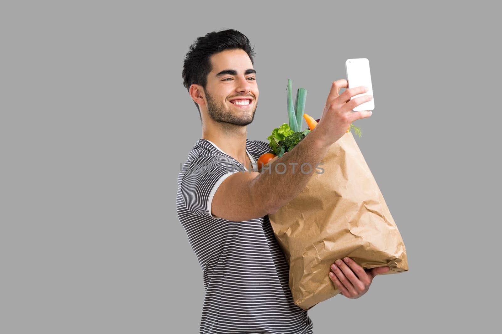 Handsome young man carrying a bag full of vegetables and making a selfie, isolated over gray background