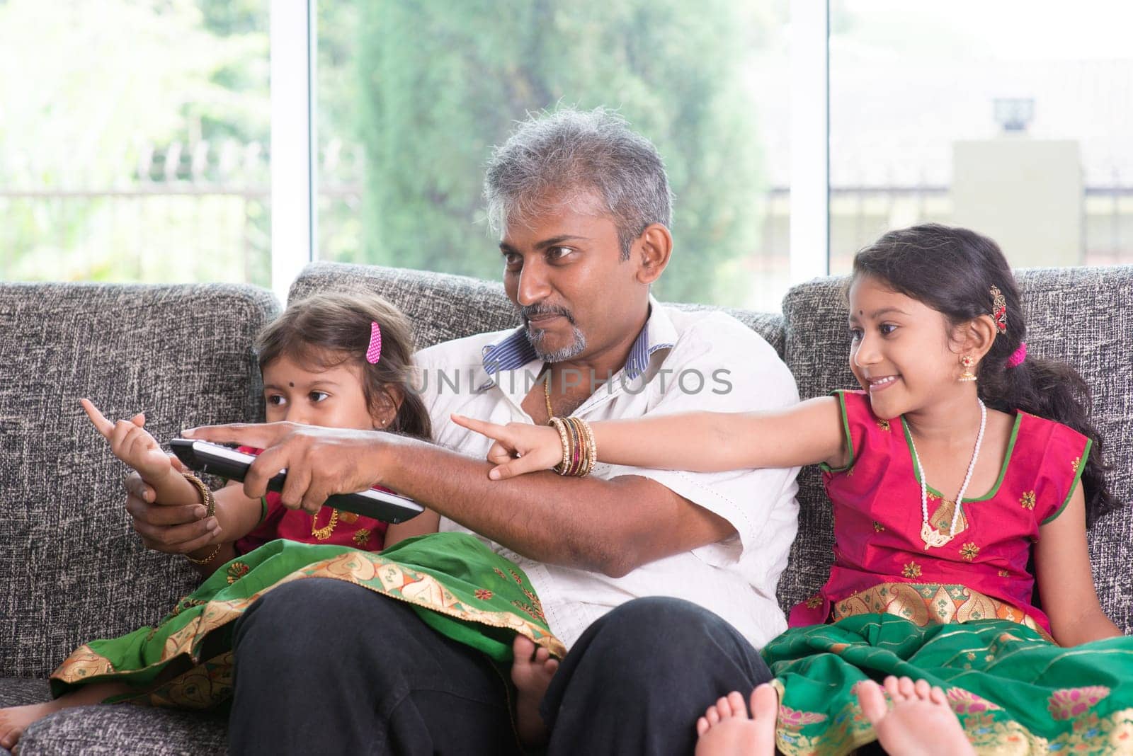 Happy Indian family watching tv together, switching channel with remote control, father and children indoor living lifestyle at home.