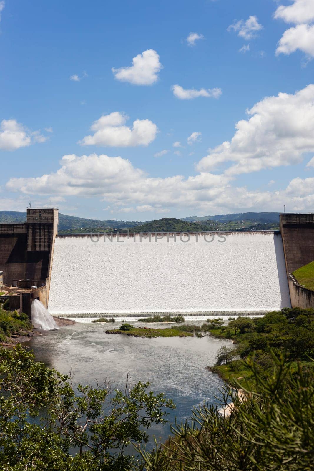 Dam wall full flowing river water over weir wall into summer valley.