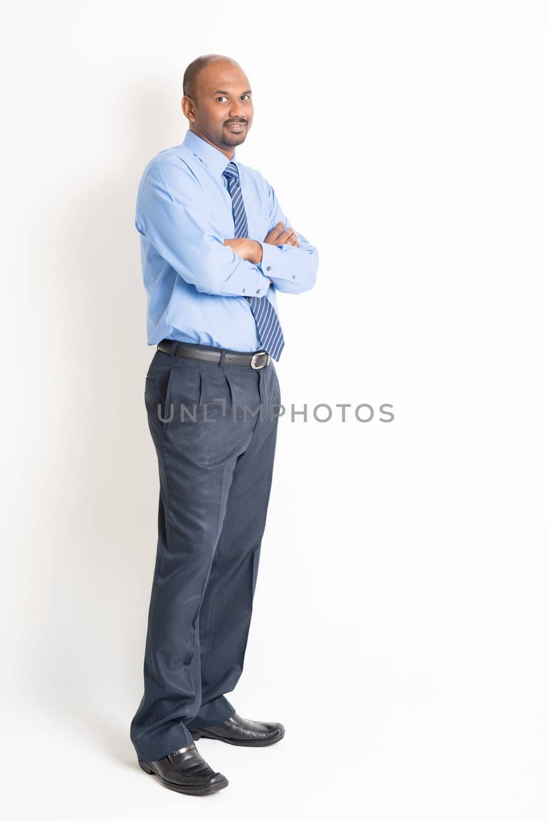 Portrait of full body mature Indian business man with blue shirt arms crossed standing on plain background.